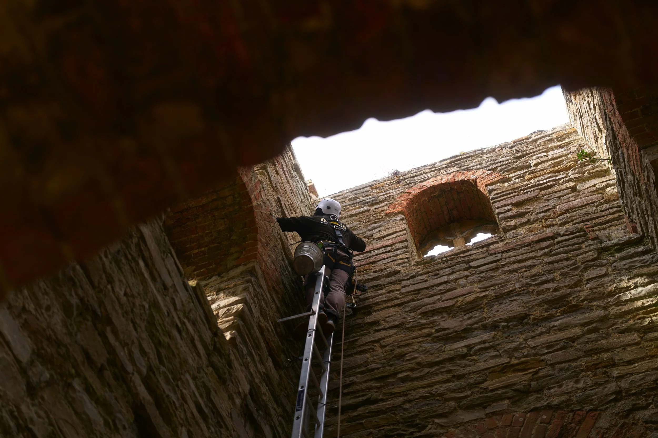 A person wearing a helmet and climbing gear ascending a ladder inside an old brick tower or structure with windows, looking up towards the open sky.