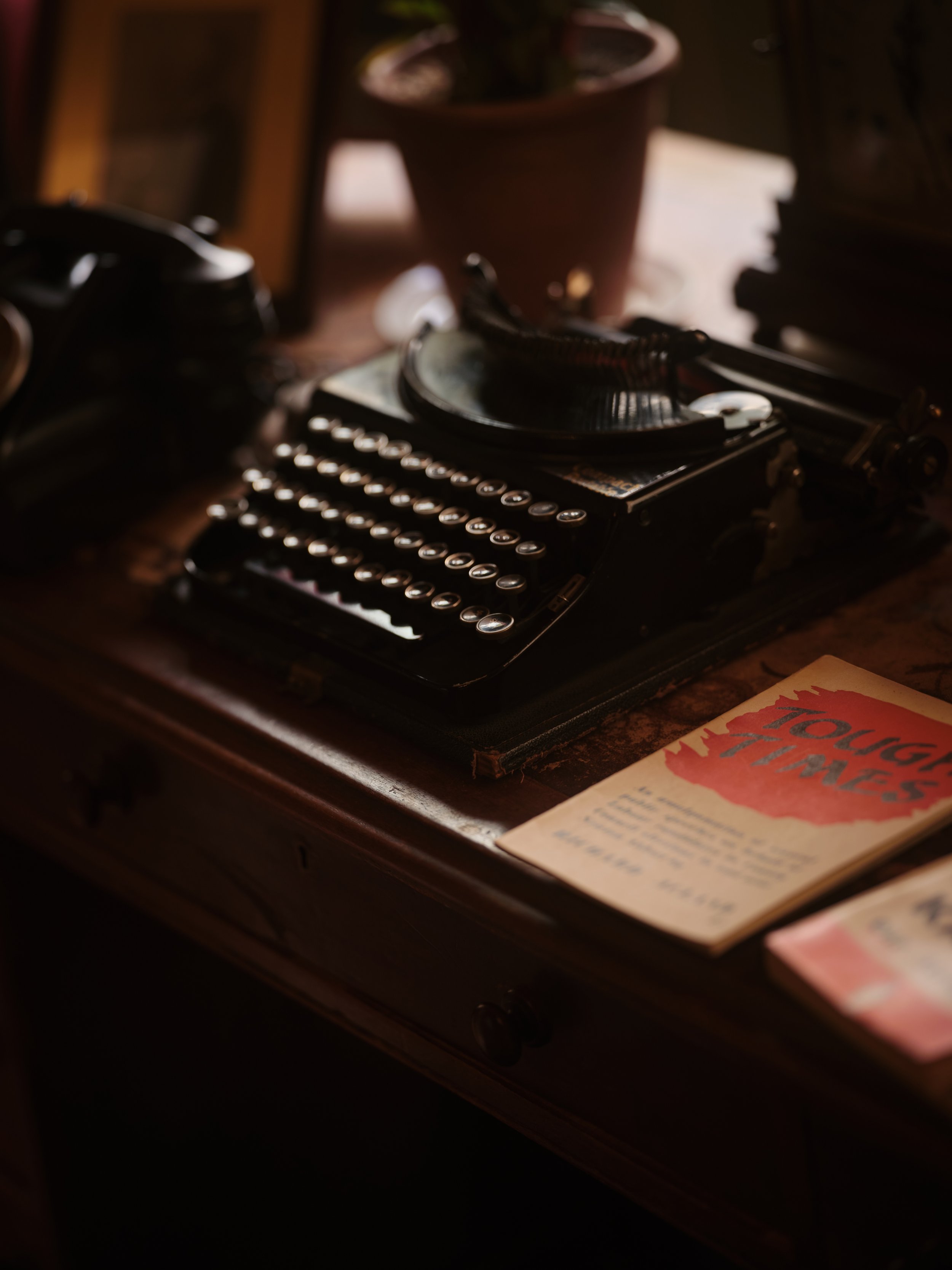 A vintage typewriter with round keys sitting on a wooden desk. There is a potted plant in the background and a partially visible booklet or paper on the desk.