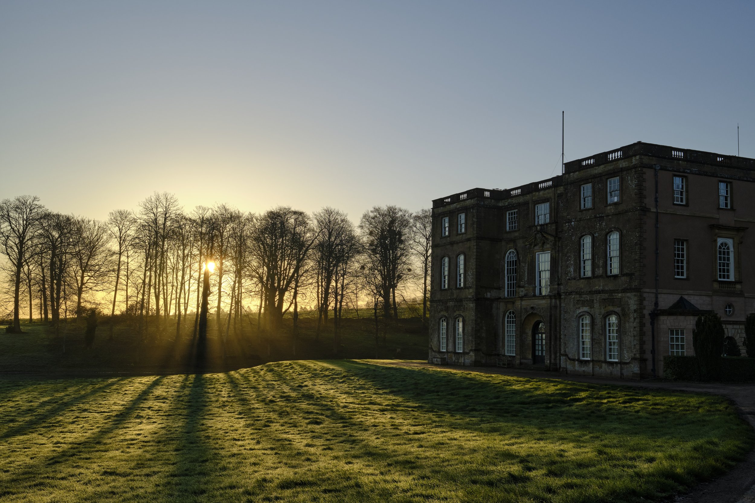 Copyright Laurence Cummins Photography / Halswell Park - Sunset over a grassy lawn with long shadows, a large historic building on the right, and leafless trees in the background.