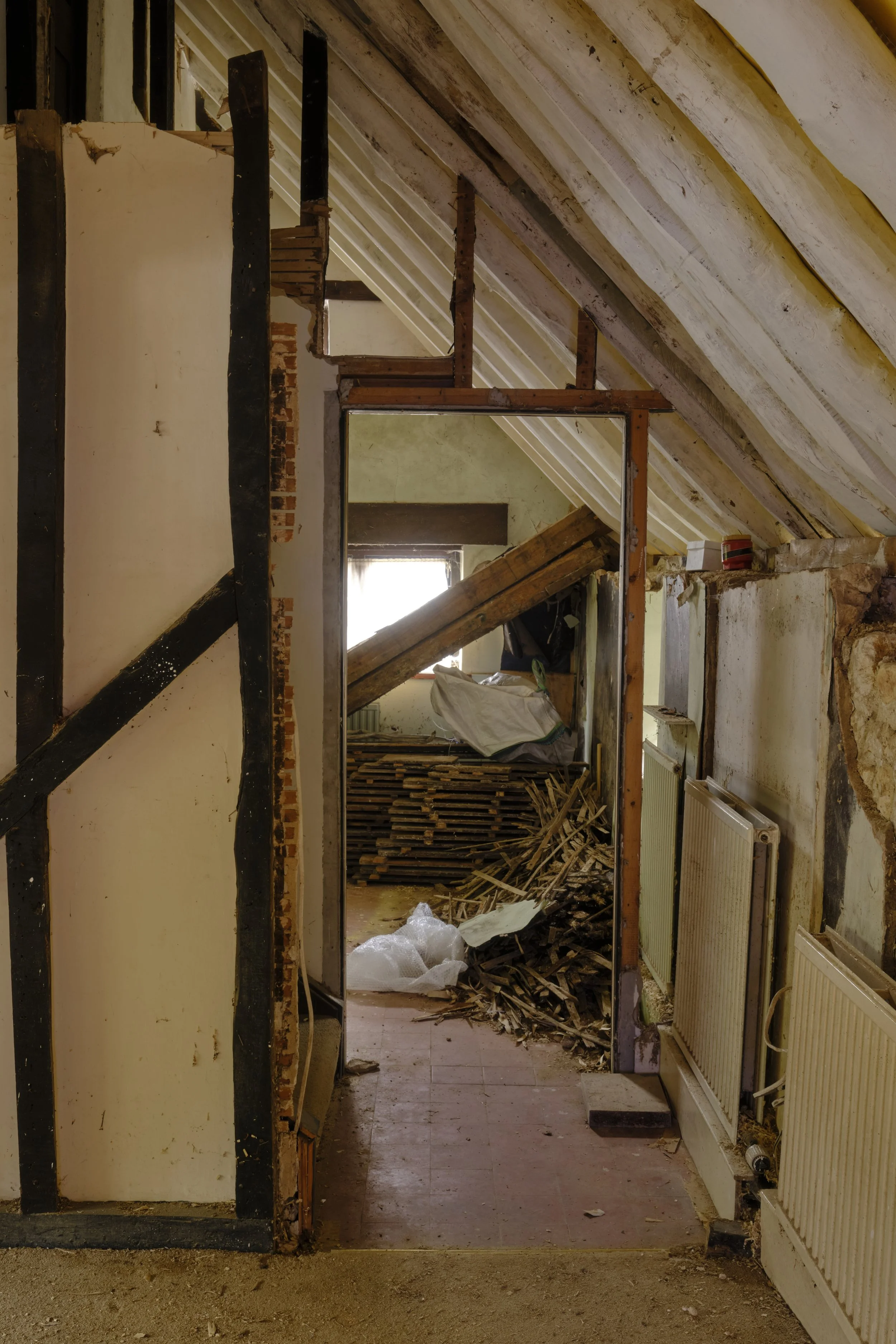 An interior of a house under renovation with exposed wooden beams, broken walls, and construction materials scattered on the floor.