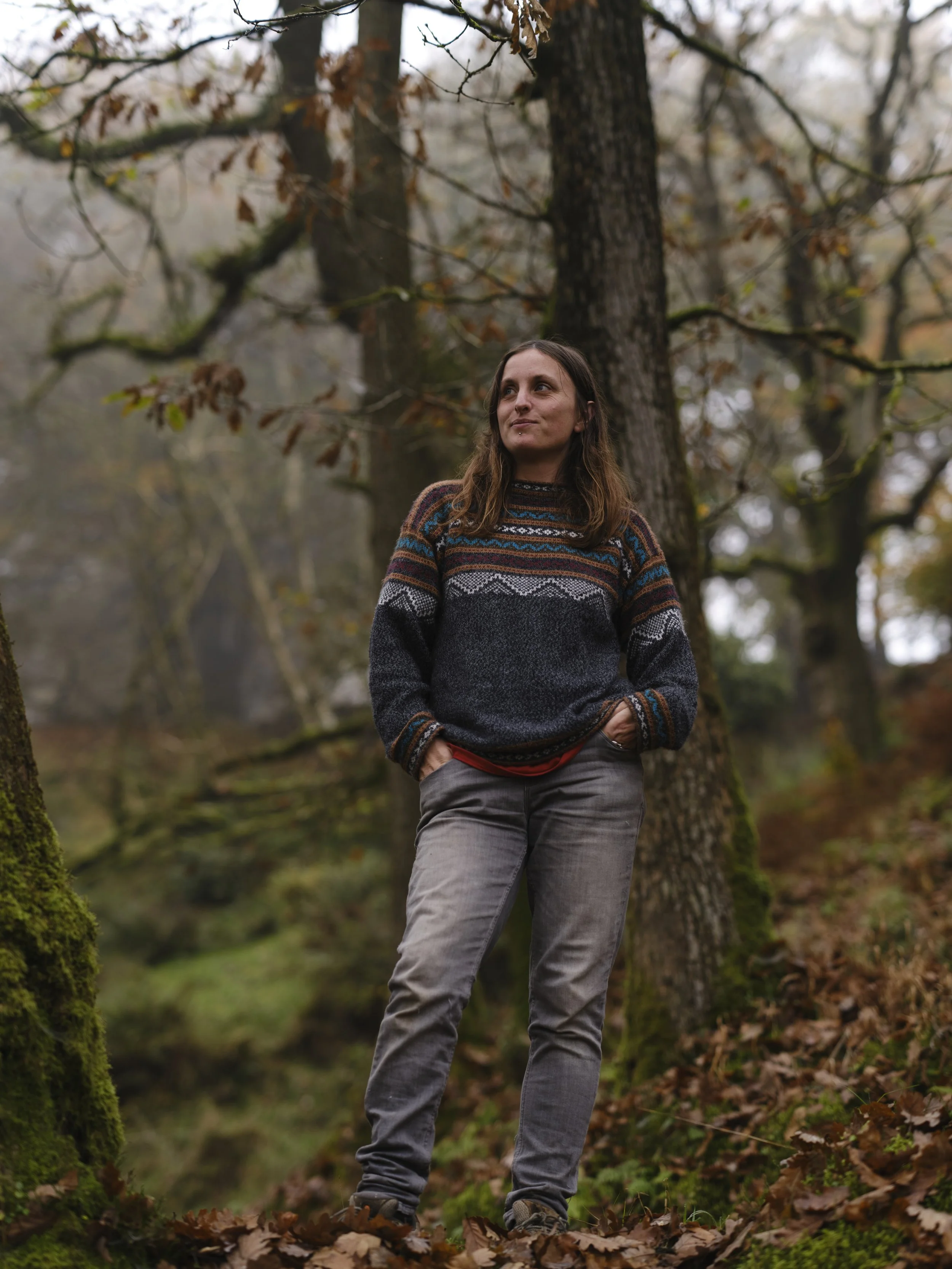 Copyright Laurence Cummins Photography - Woman standing in a forested area with trees and fallen leaves, wearing a multicolored sweater and gray jeans.