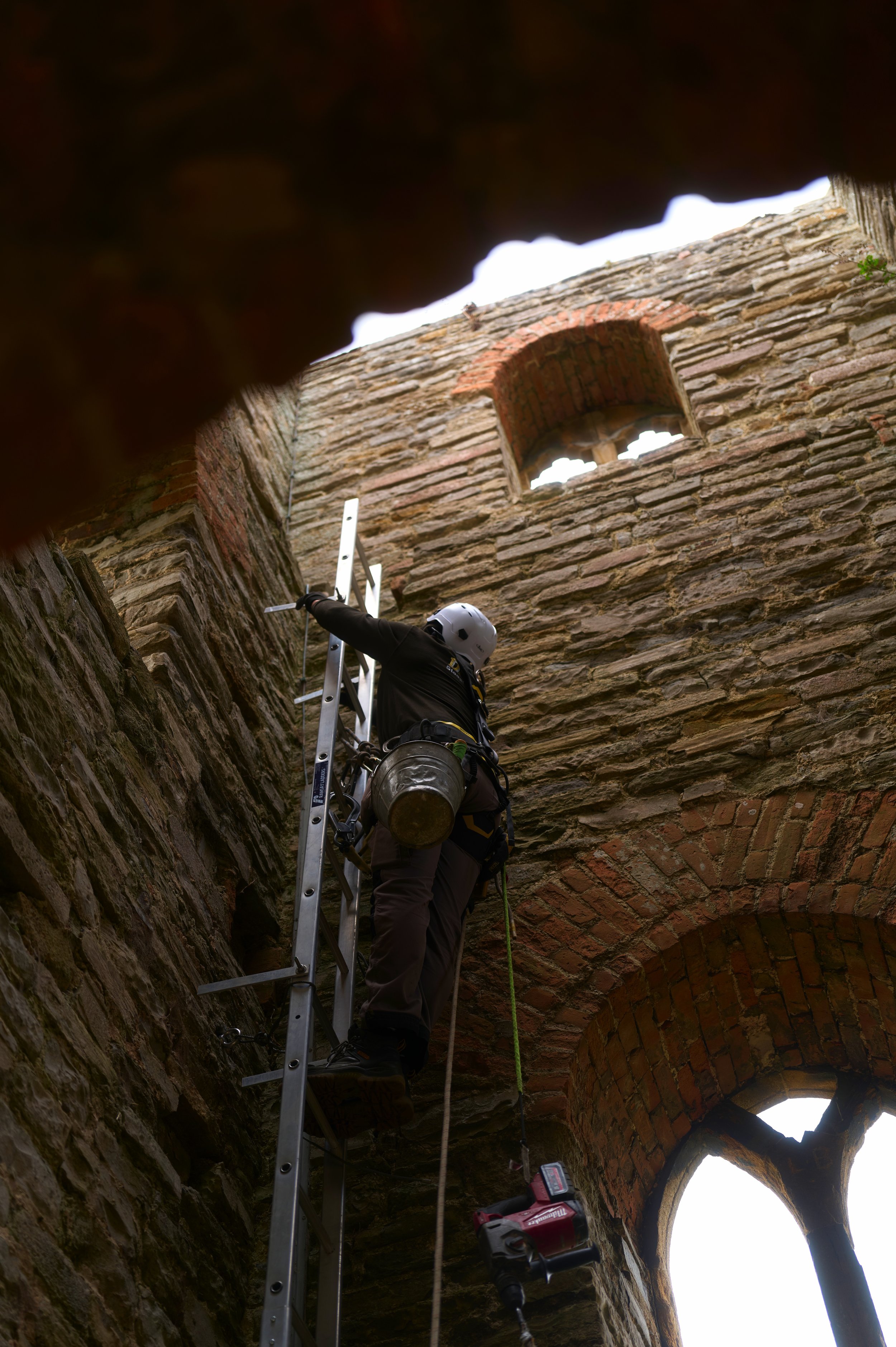 A worker standing on a ladder and inspecting the brick and stone structure of a historic building in daylight. The worker is wearing a safety helmet and harness, with tools nearby.