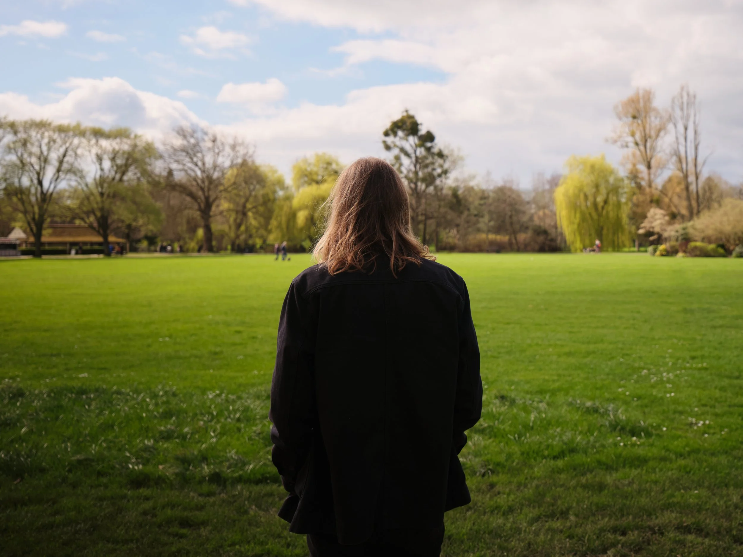 A person with shoulder-length brown hair, wearing a black jacket, standing in a green park with trees and cloudy sky in the background.