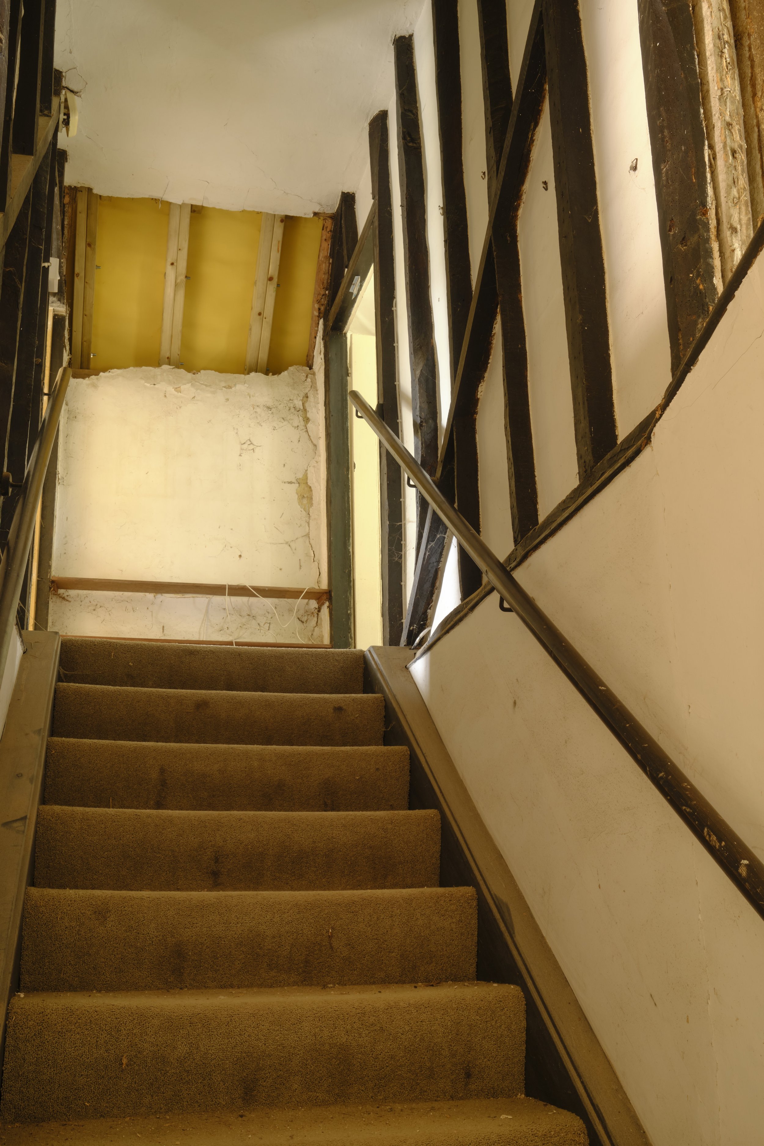 A residential staircase under renovation with carpeted stairs, a wooden framework, exposed wall and ceiling studs, and construction materials.