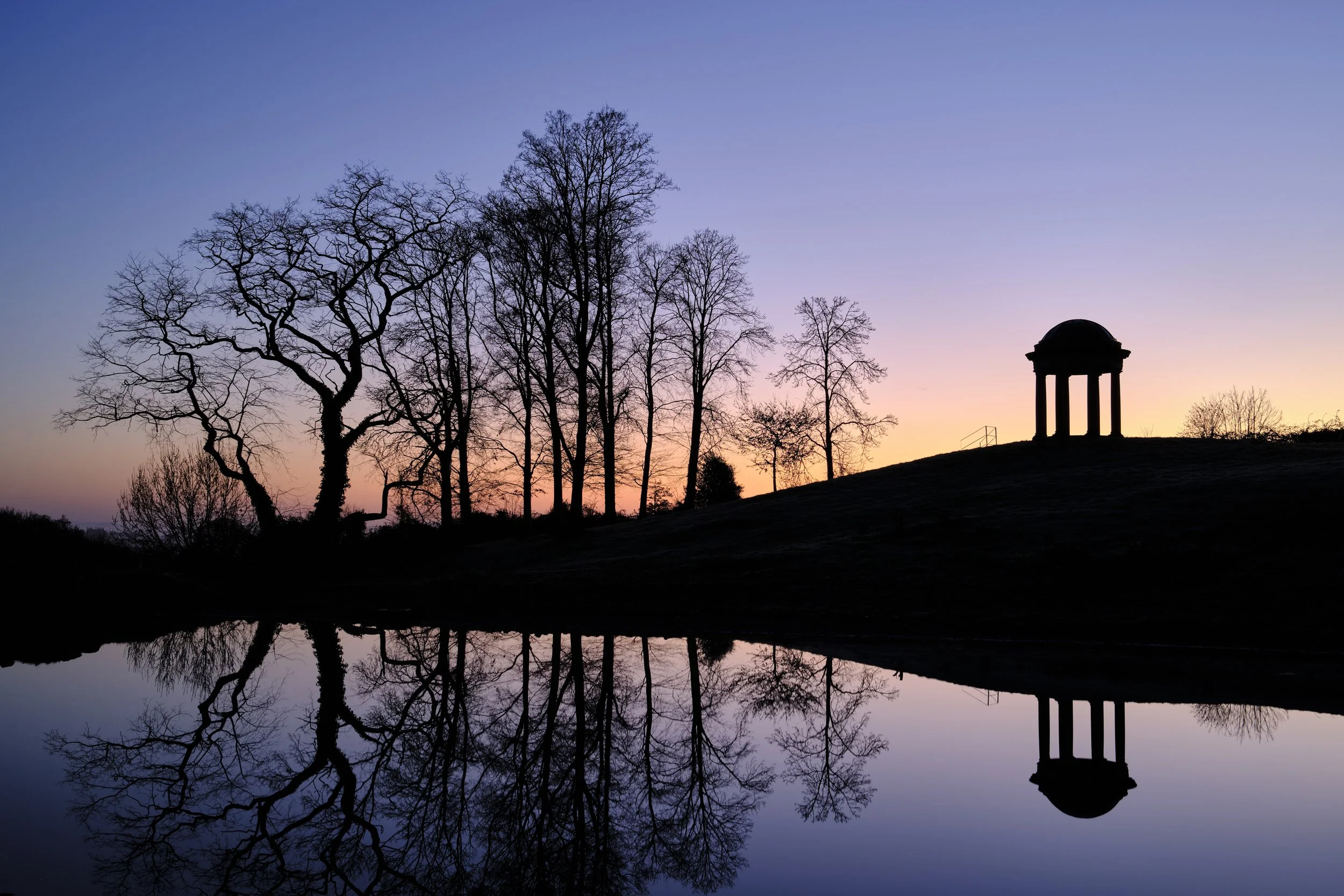 Copyright Laurence Cummins Photography / Halswell Park - Silhouetted trees and a small pavilion on a hill at sunset, with their reflection in a body of water.
