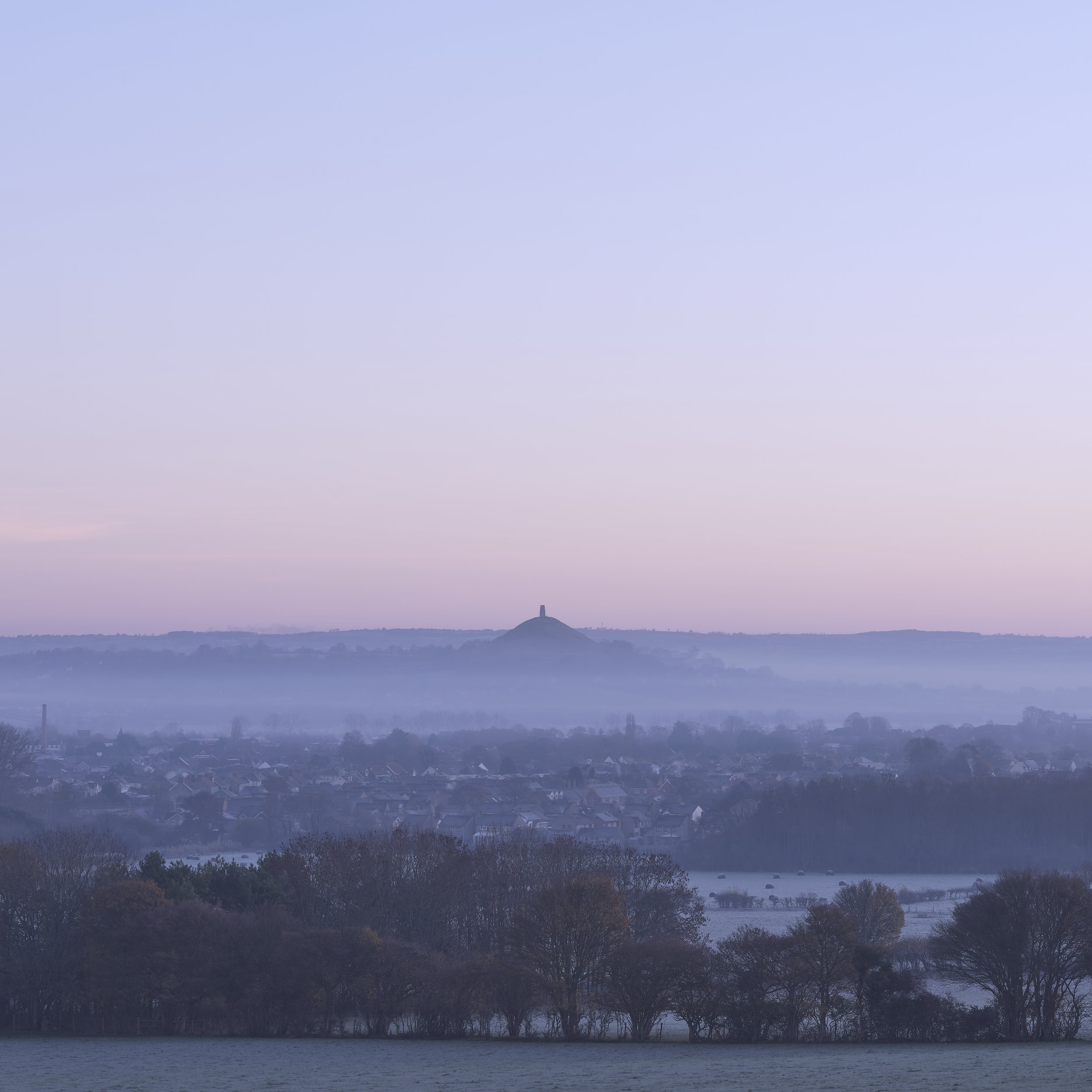 Copyright Laurence Cummins Photography - Landscape view of a town with a hill in the distance, topped with a monument or structure, surrounded by trees and misty hills at dawn or dusk.