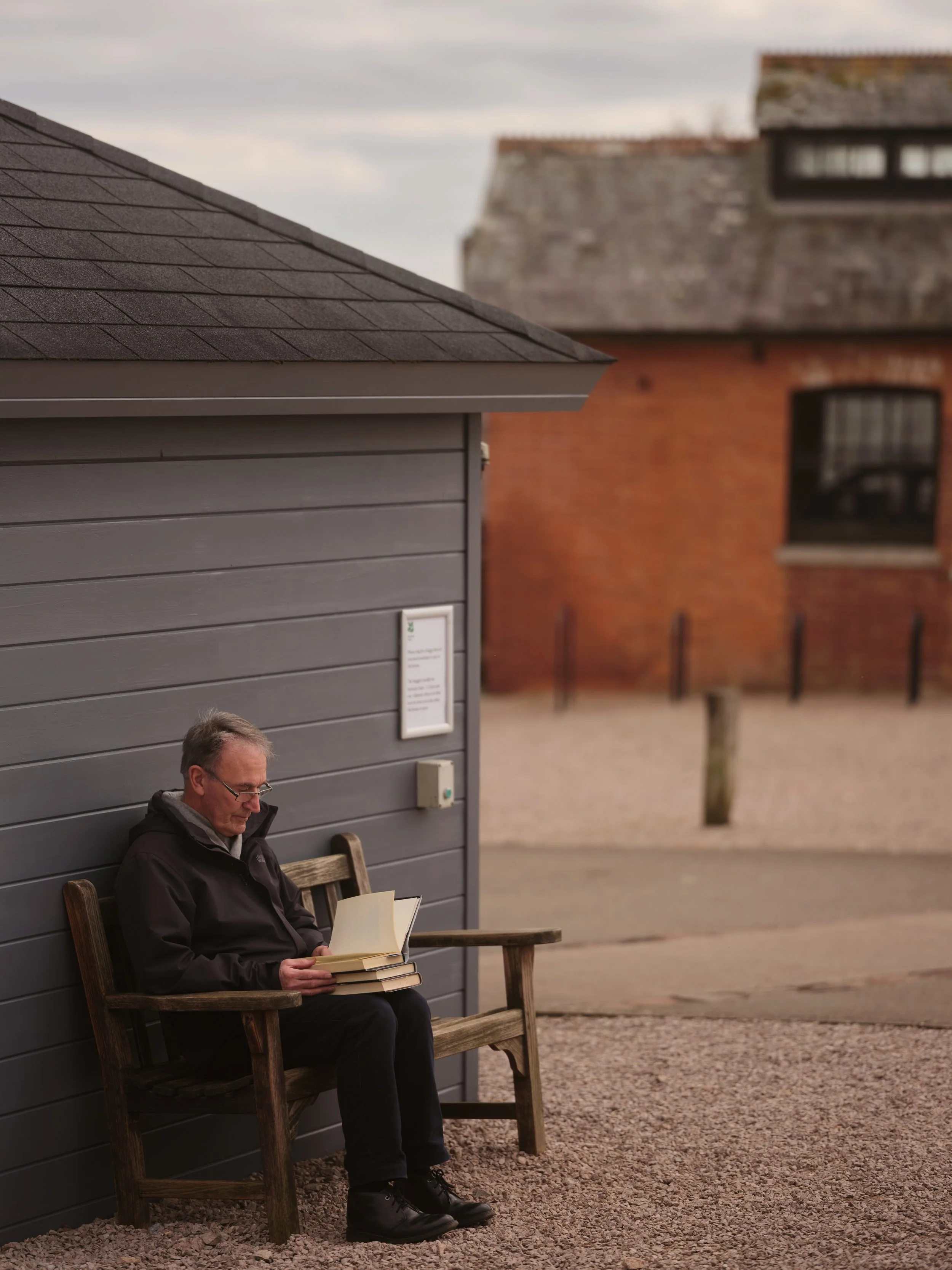 A man with glasses sitting on a wooden bench outside a gray building, reading a book. The building has horizontal siding, and there is a framed notice or sign on it. In the background, there are other buildings with red brick walls and a gravel groun