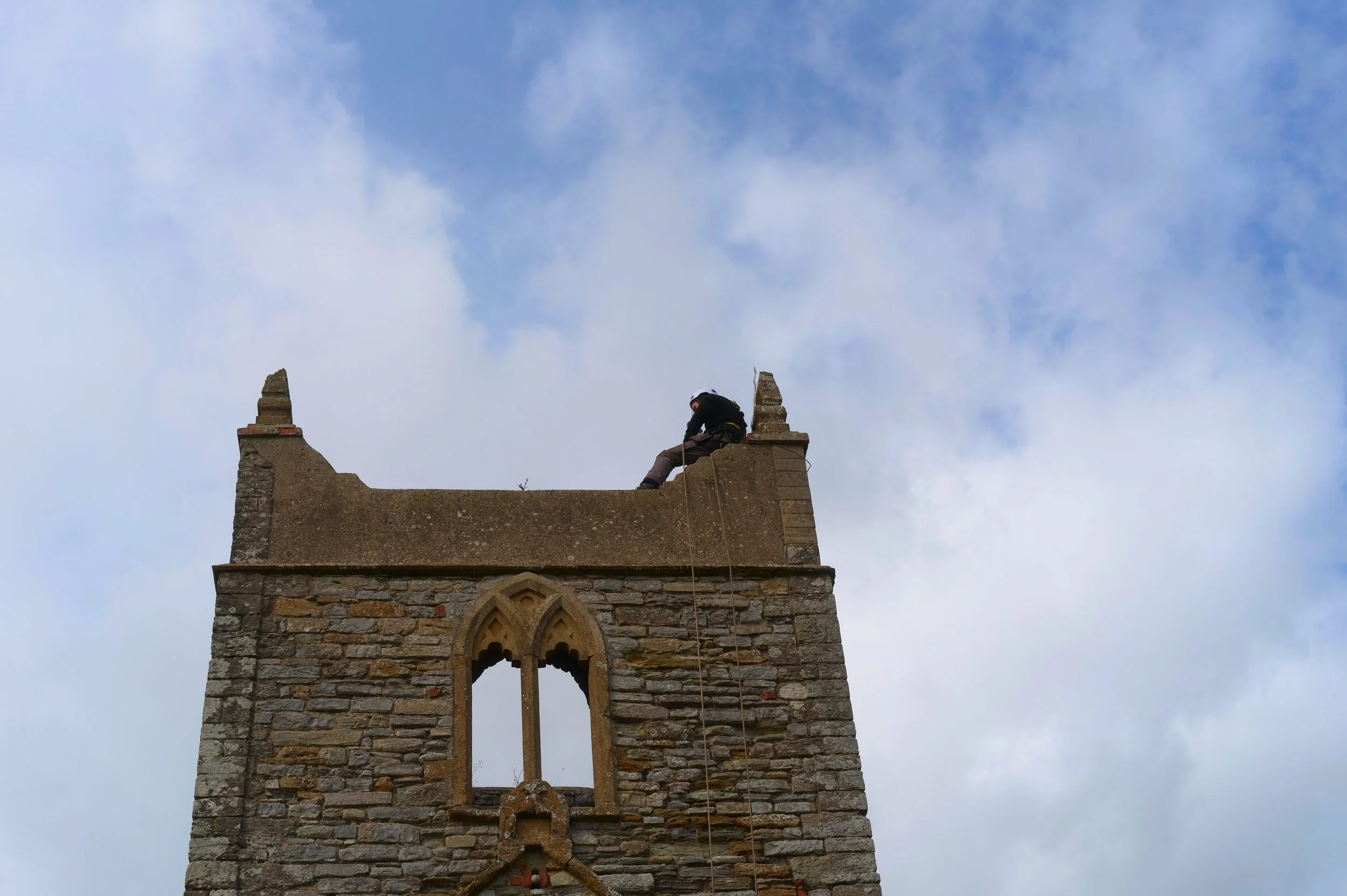 A person sits on the edge of a stone tower or church steeple, wearing a helmet and harness, with a cloudy sky in the background.