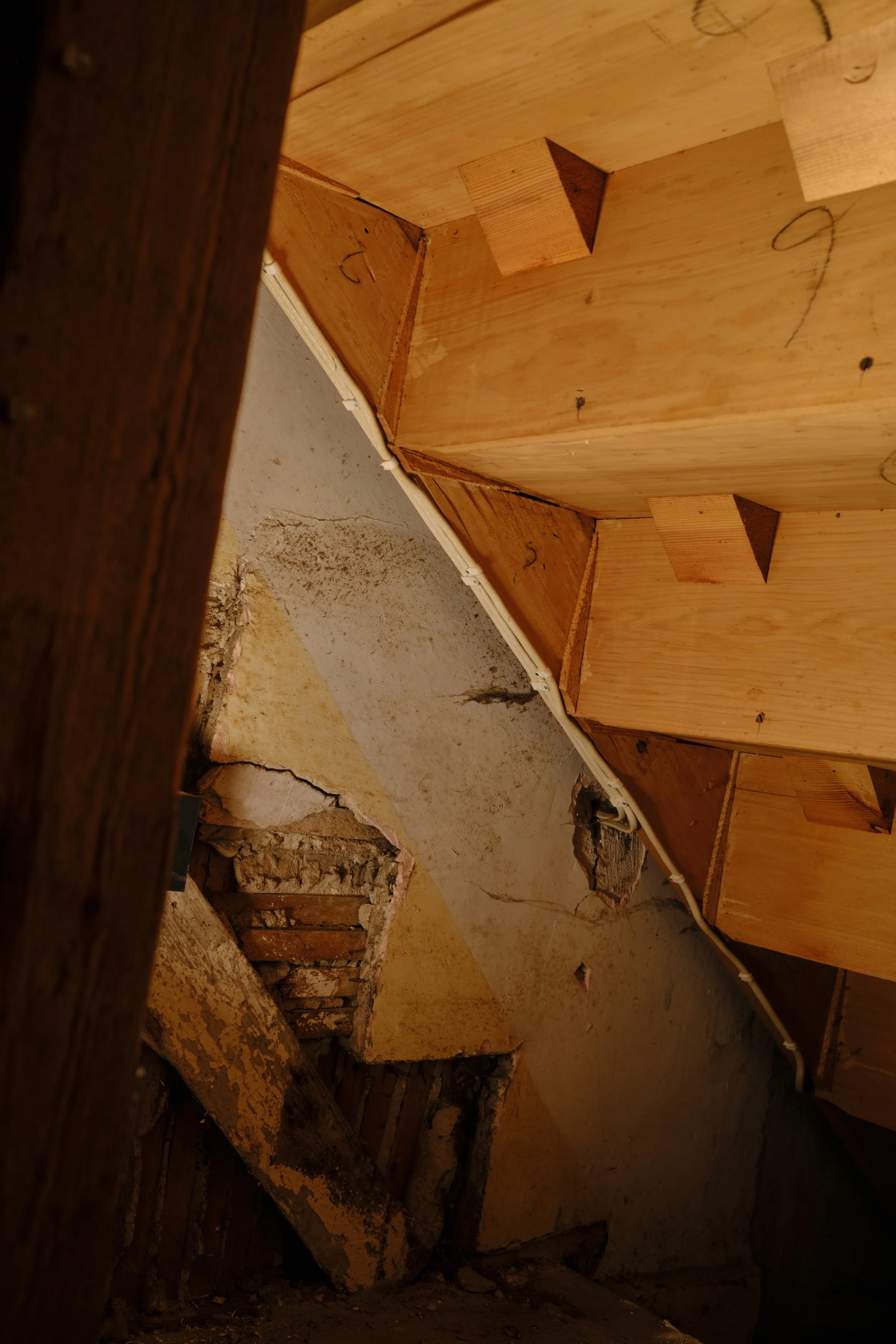 Ceiling with exposed wooden beams and partially damaged insulation, showing a hole in the wall below with exposed brick and insulation.