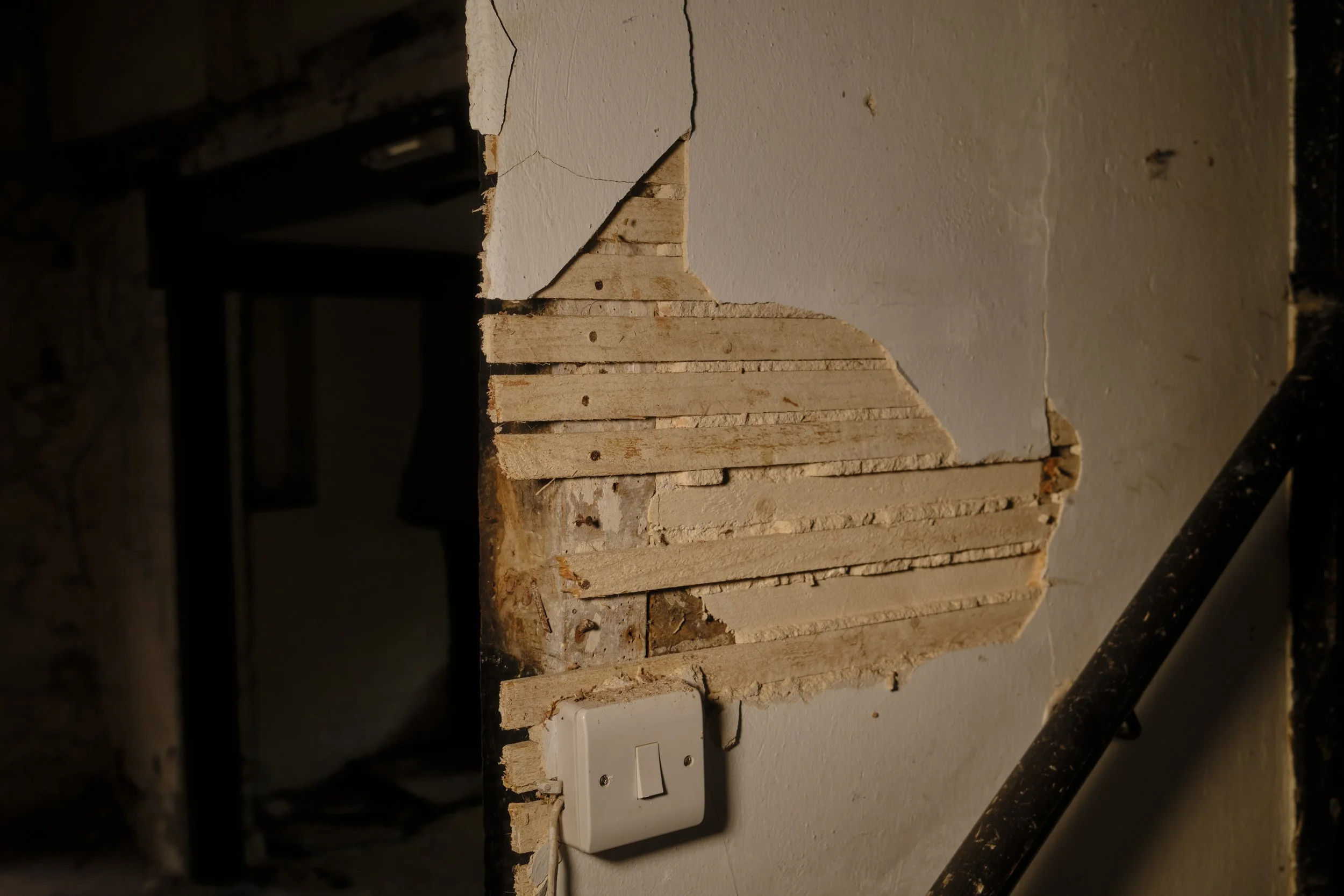 Close-up of a wall with a section of plaster removed, exposing wooden laths underneath, and a white electrical switch at the bottom.
