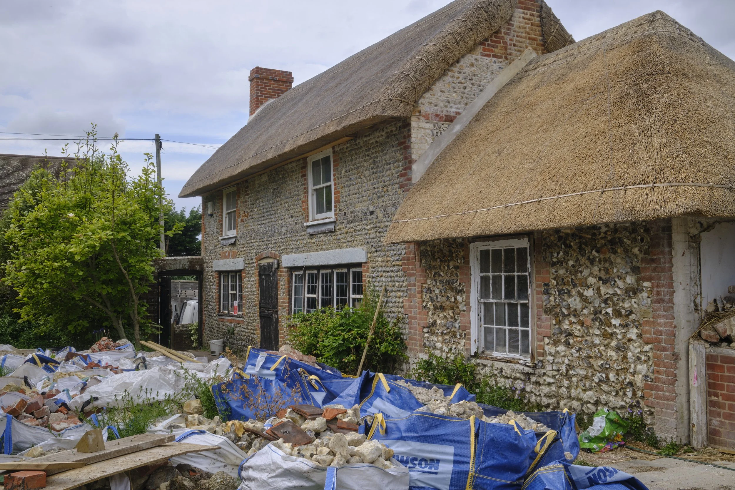 A house with a thatched roof and stone brick walls under renovation or repair, with construction debris including bricks, rubble, and a blue bag with building materials in the foreground.