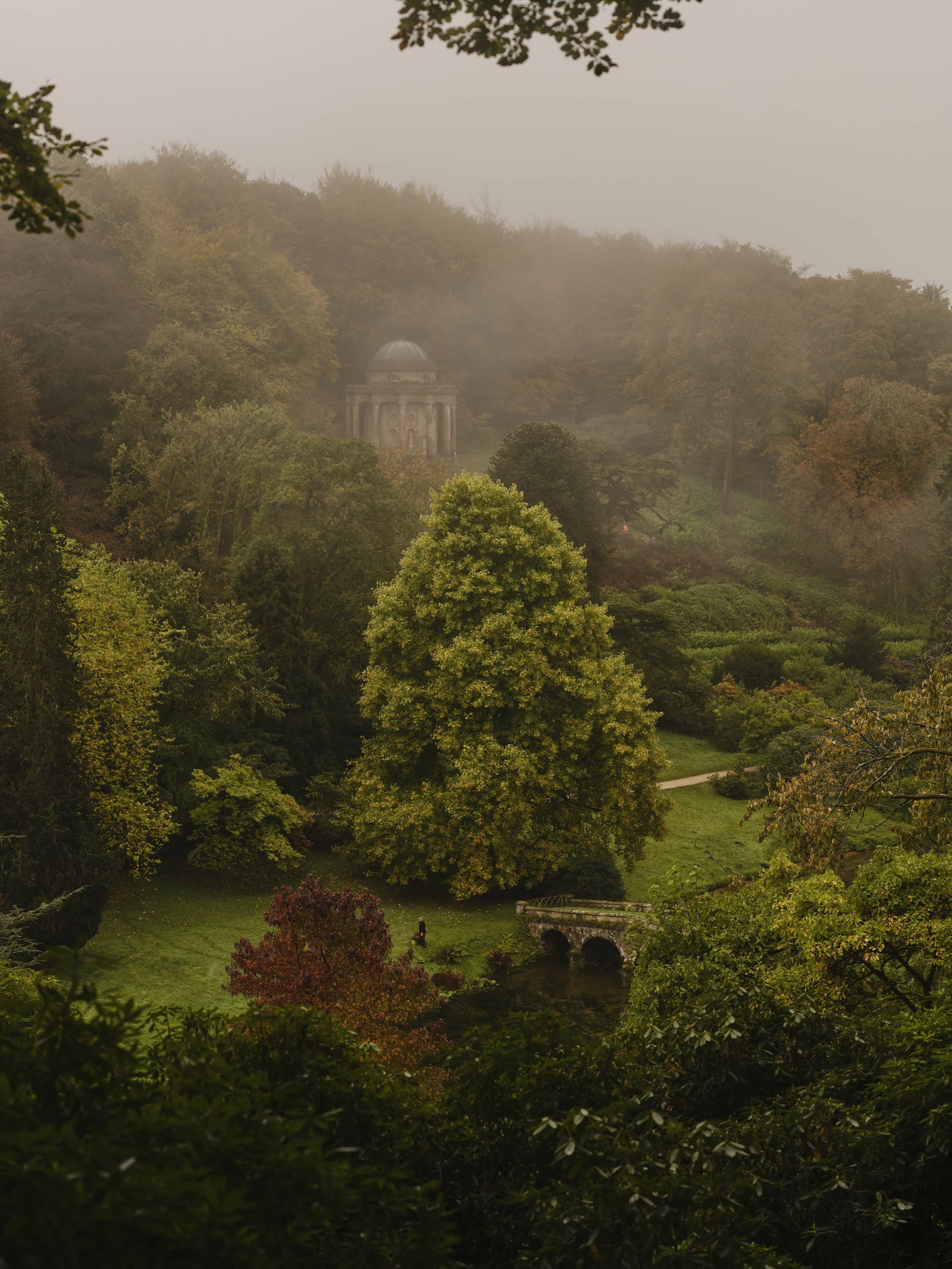 A foggy, wooded landscape with a classical building with a dome in the background, surrounded by various trees and a small stone bridge over a stream.