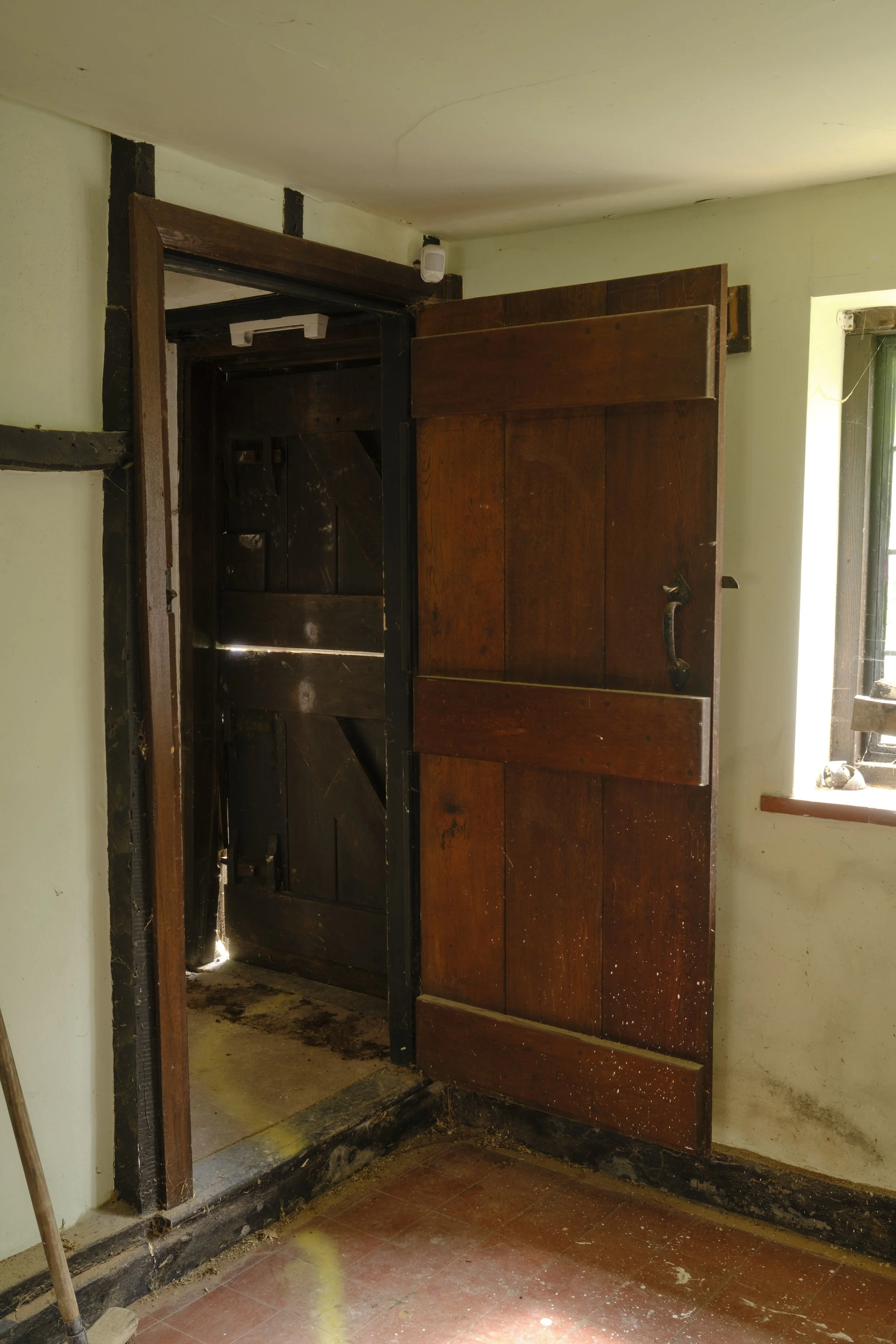 A partially removed wooden door frame with an open dark wooden door, revealing an interior space with dark wood paneling and a doorway. The area shows signs of damage and dirt on the floor and walls, with a small window on the right side of the image