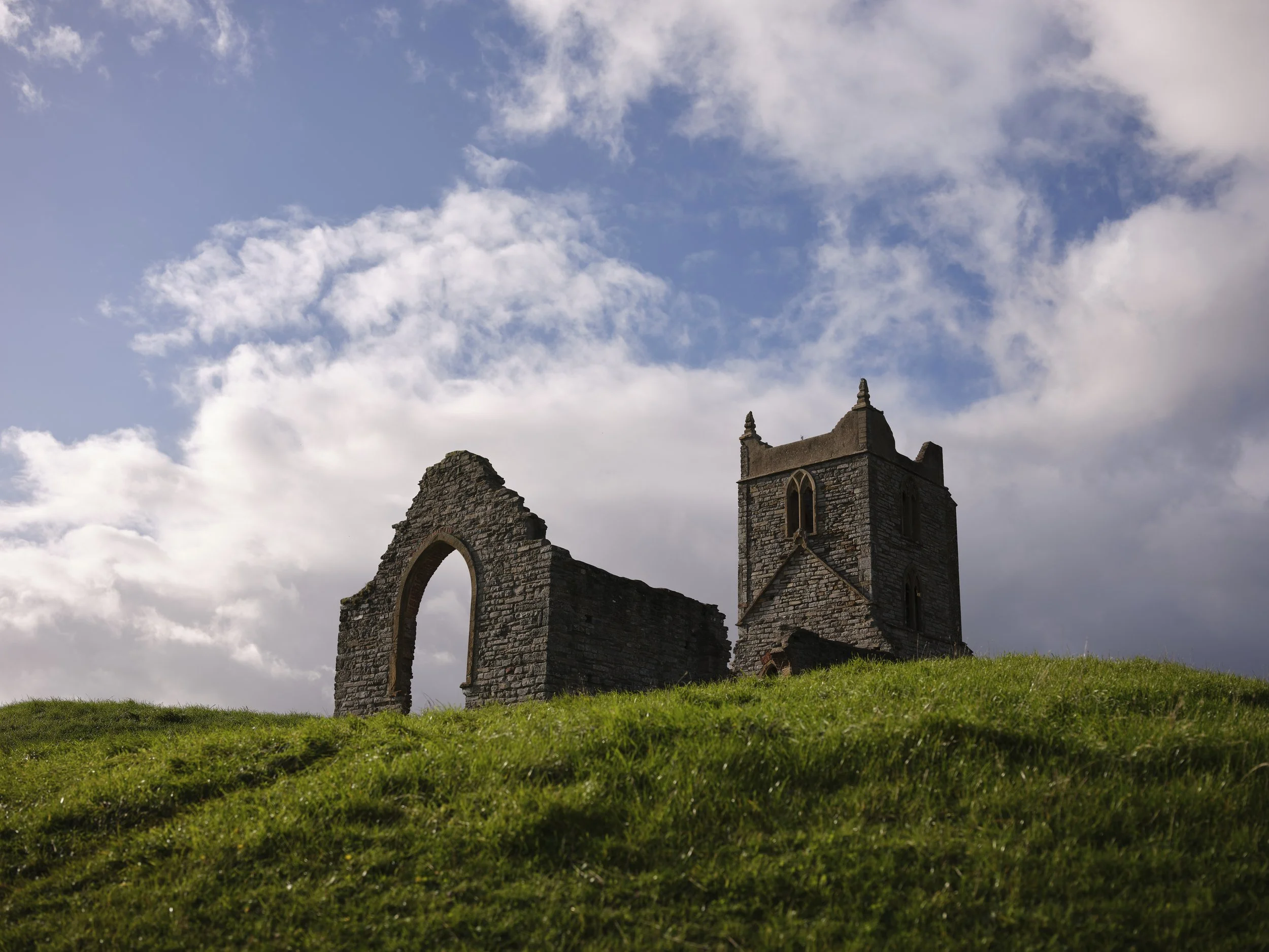 Copyright Laurence Cummins Photography - Ruins of an old stone church with a tower, situated on a grassy hill under a partly cloudy sky.