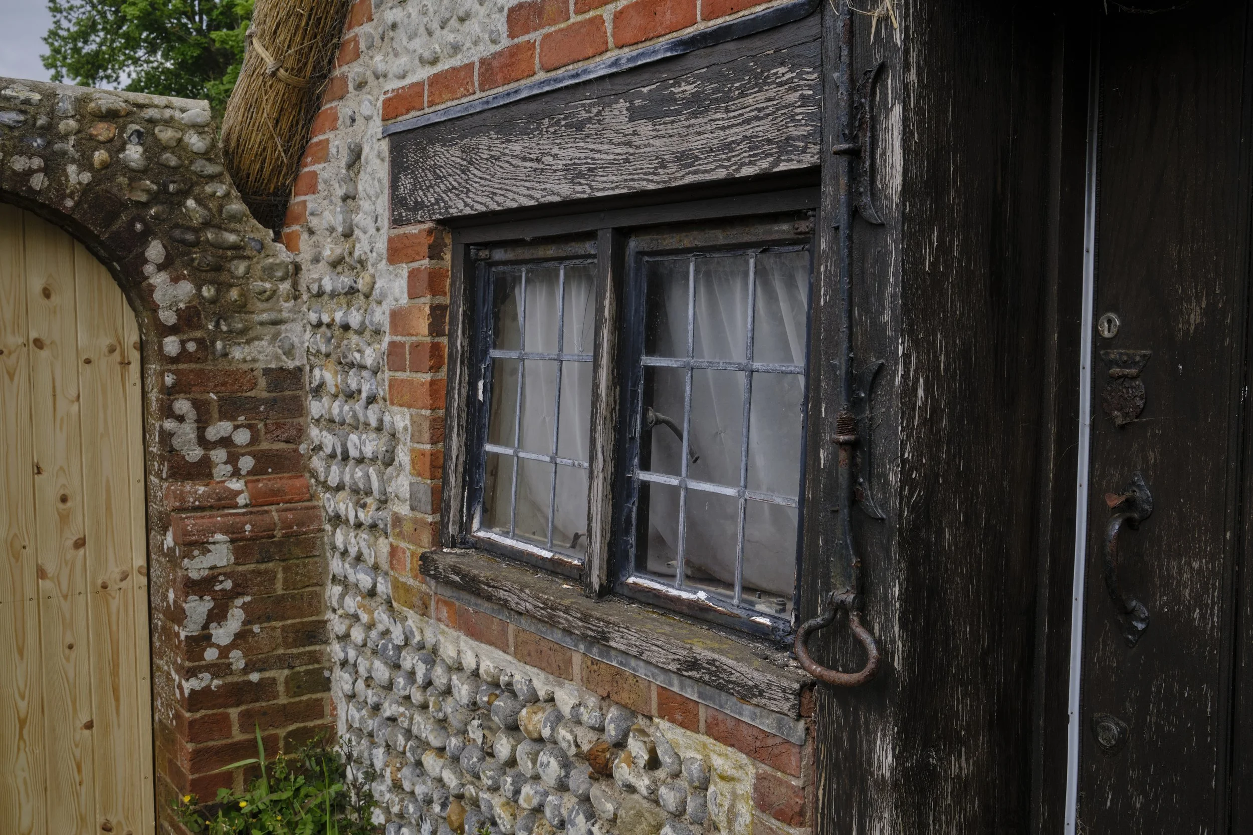 A close-up of an exterior wall showcasing a combination of materials, including weathered dark wood, red brick, and pebbled stones, with an old window with curtained panes and some garden plants at the bottom.