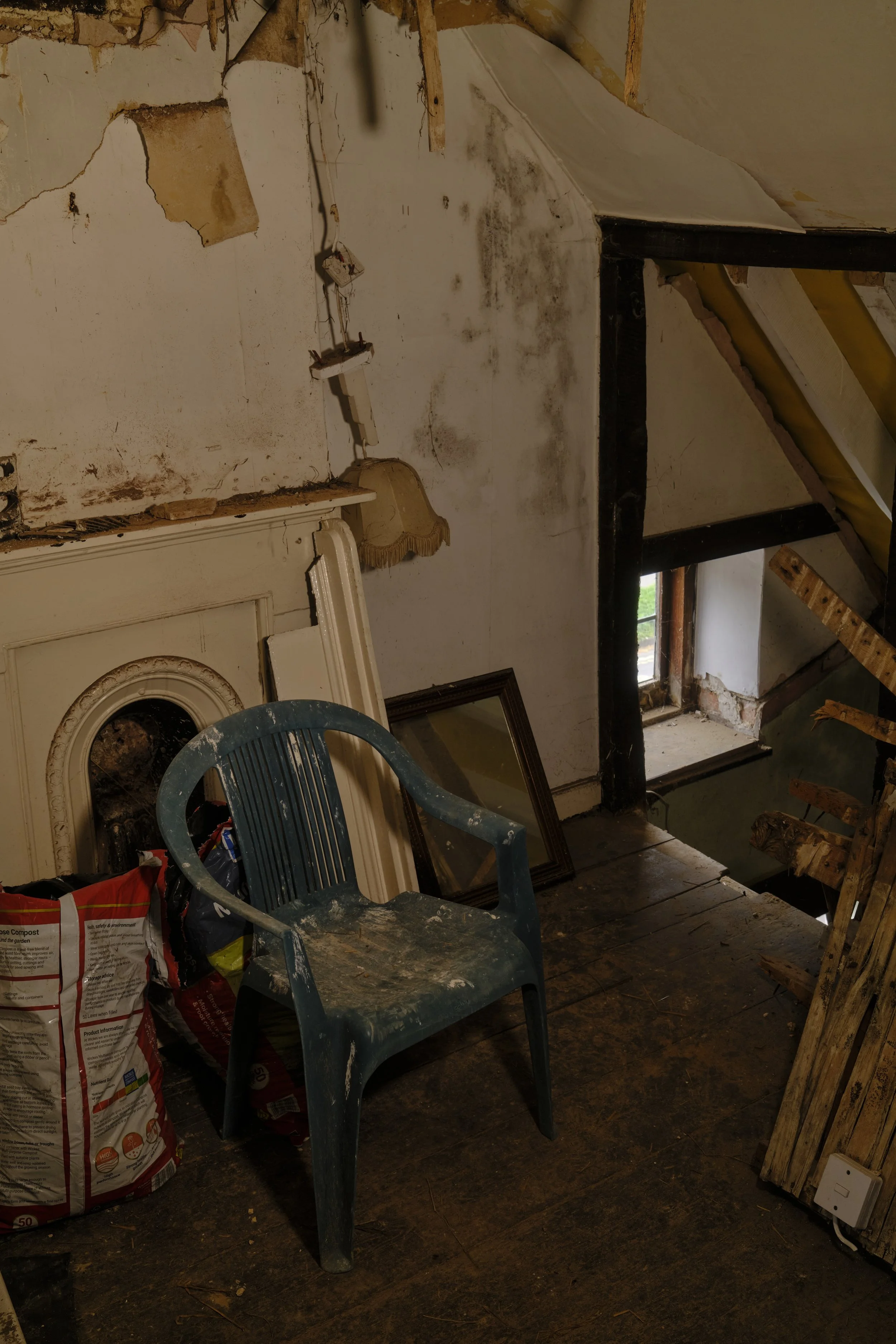 A room under renovation with exposed walls and ceiling, a plastic chair covered in dust, a bag of compost, an old mirror, and various construction materials scattered around. There is a small window letting in some natural light.