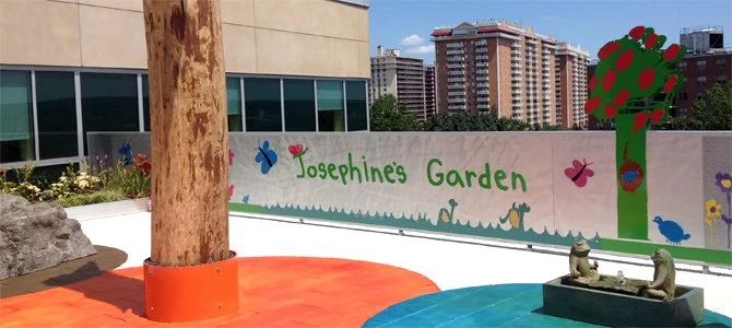 Colorful children's mural titled "Josephine's Garden" with drawings of flowers, butterflies, a tree, and animals, featuring a small frog statue in front.
