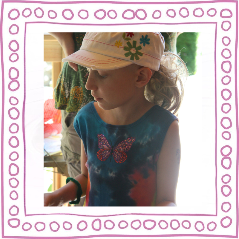A young girl with curly blonde hair wearing a tie-dye tank top with a butterfly design and a pink hat with floral embroidery, looking down thoughtfully.