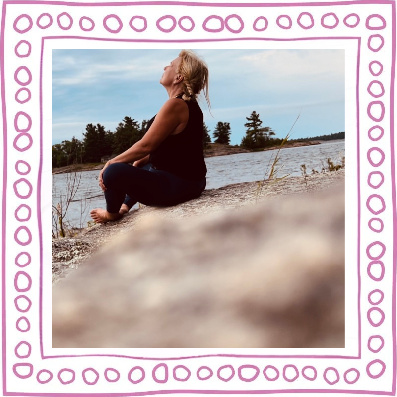 A woman sitting on a rock by a lake, looking upward toward the sky, with trees and clouds in the background.