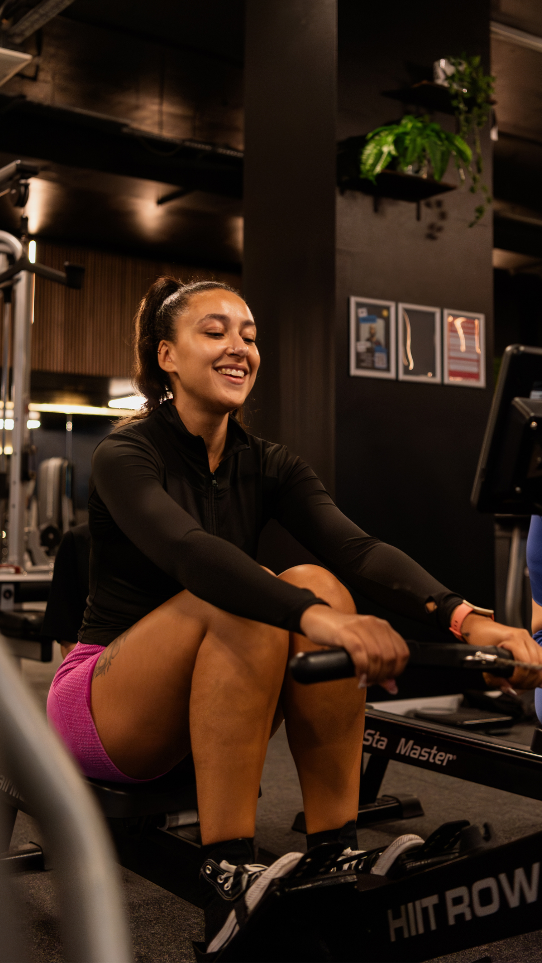 A woman with dark hair in a ponytail working out on a rowing machine in a gym. She is smiling, wearing a black long-sleeve top and pink shorts, with a watch on her wrist.
