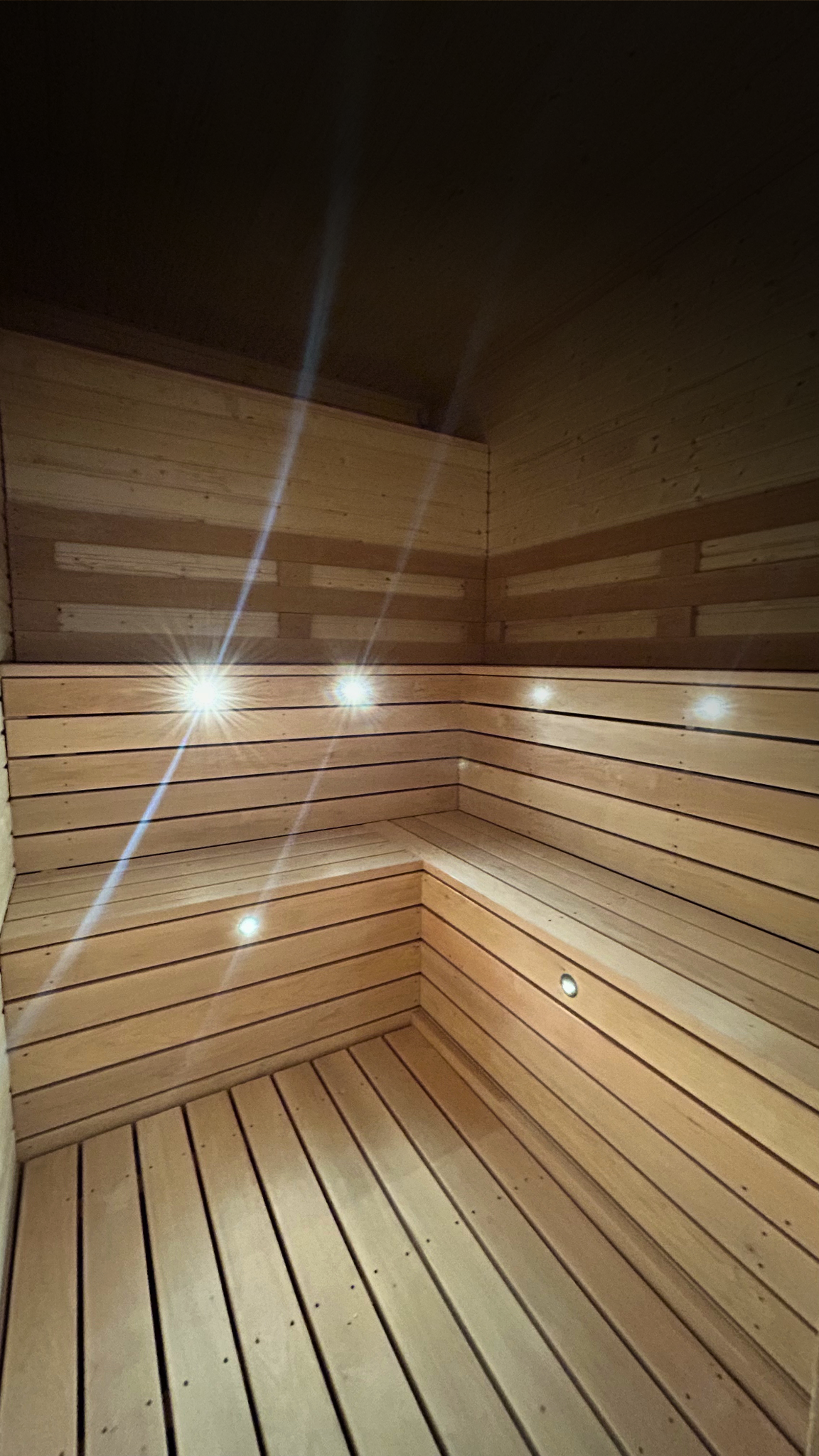 Interior of a wooden sauna with horizontal wood paneling on the walls and ceiling, and slatted wooden benches, with built-in small round lights.