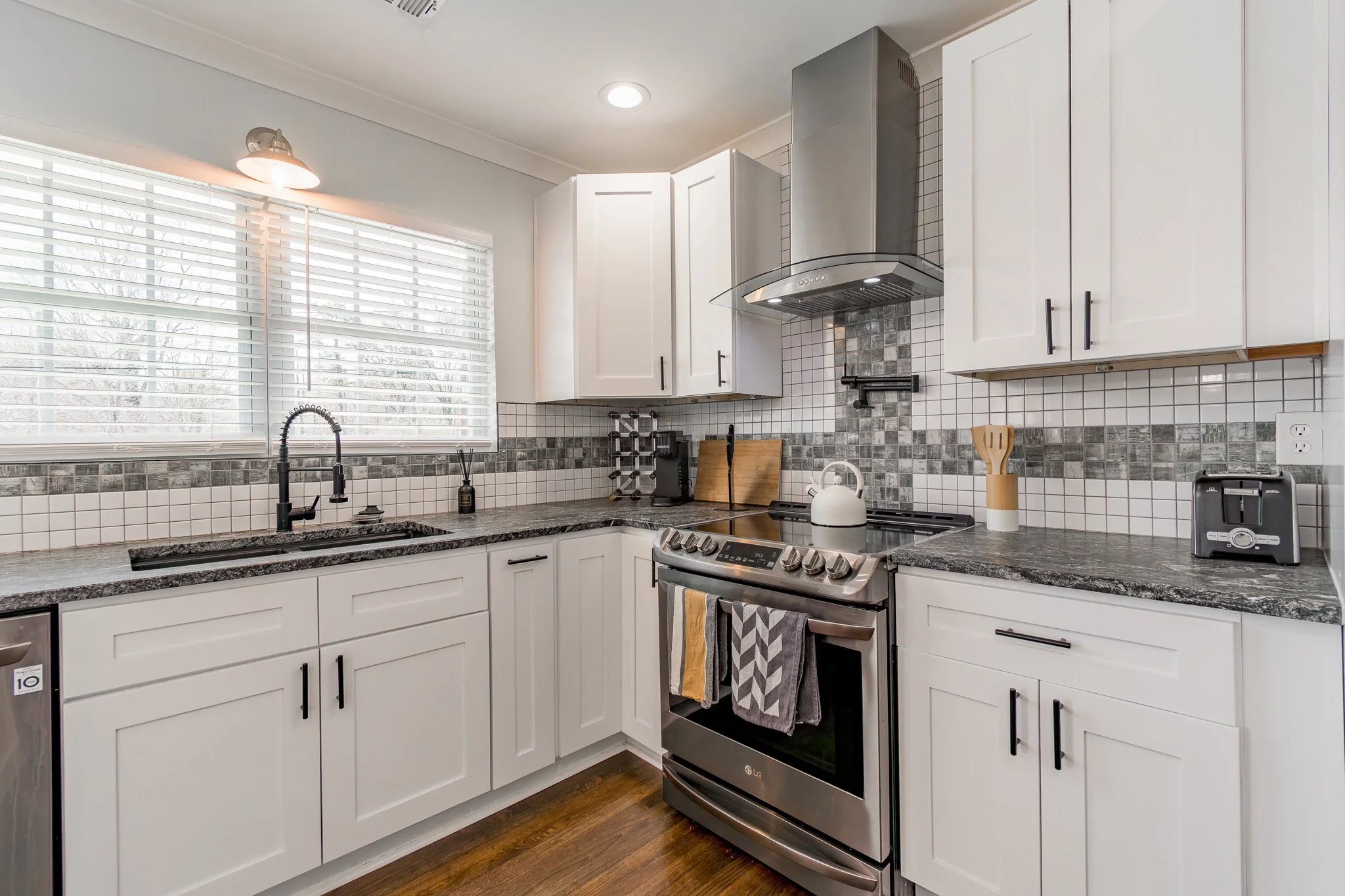 Kitchen with white cabinets, black handles, dark granite countertops, stainless steel stove, range hood, and white tiled backsplash with gray accents. There is a large window with horizontal blinds above the sink, which has a black faucet. Various ki