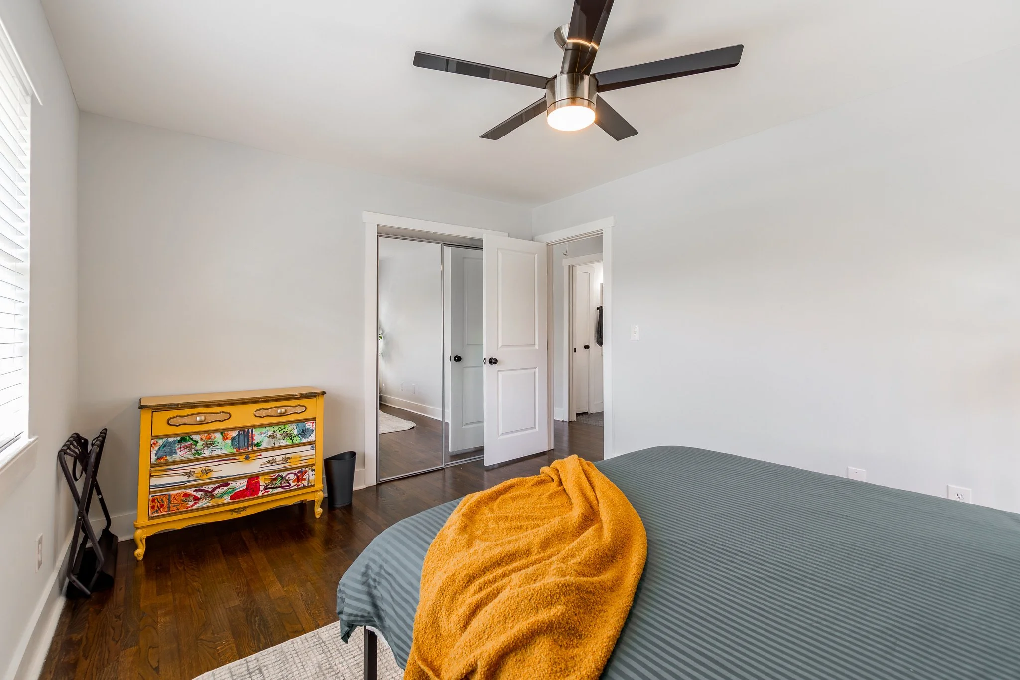 A bedroom with a bed covered in a gray striped blanket and an orange throw, a yellow dresser with colorful artwork, and a ceiling fan with six blades.