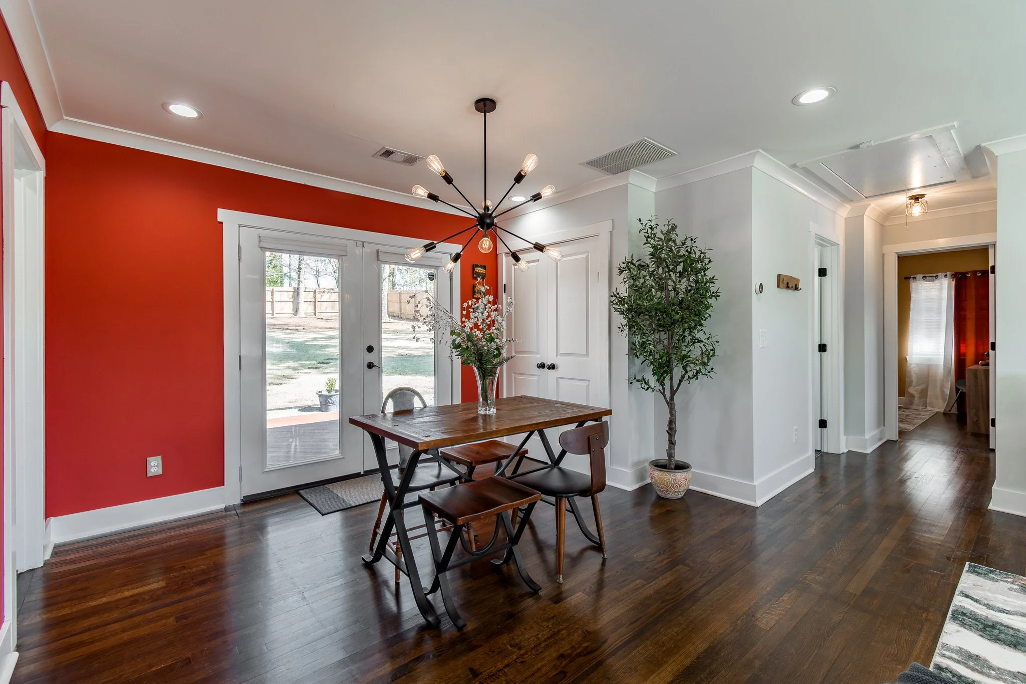 Dining room with dark wood flooring, white walls, and a red accent wall with double glass doors leading outside. A modern chandelier hangs above a wooden dining table with mixed chairs, and a potted plant is nearby.