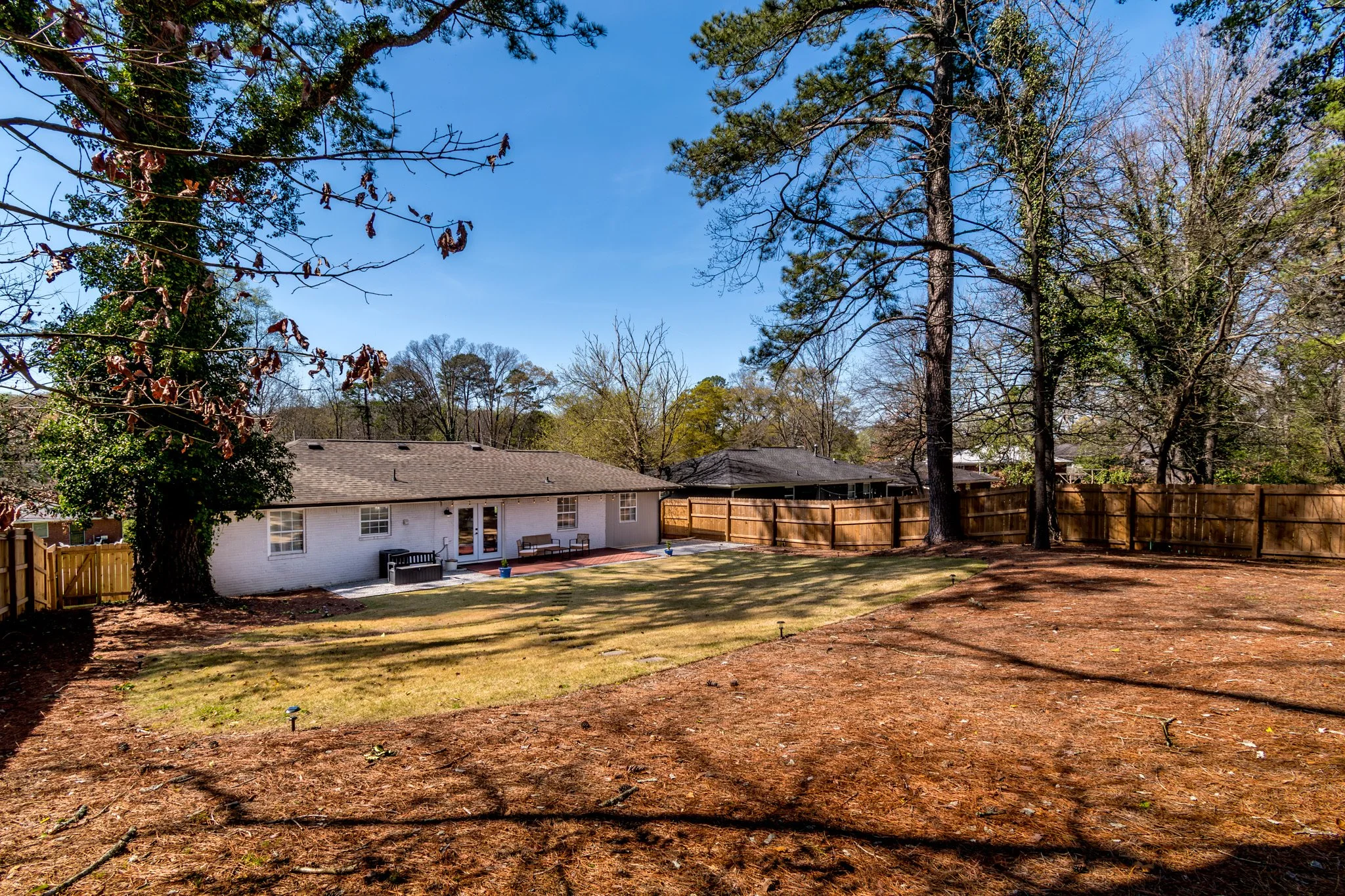 Backyard with a well-maintained lawn, a brick patio, and a white house with a gray roof. Tall trees surround the yard, and a wooden privacy fence encloses the area. The sky is clear and blue.