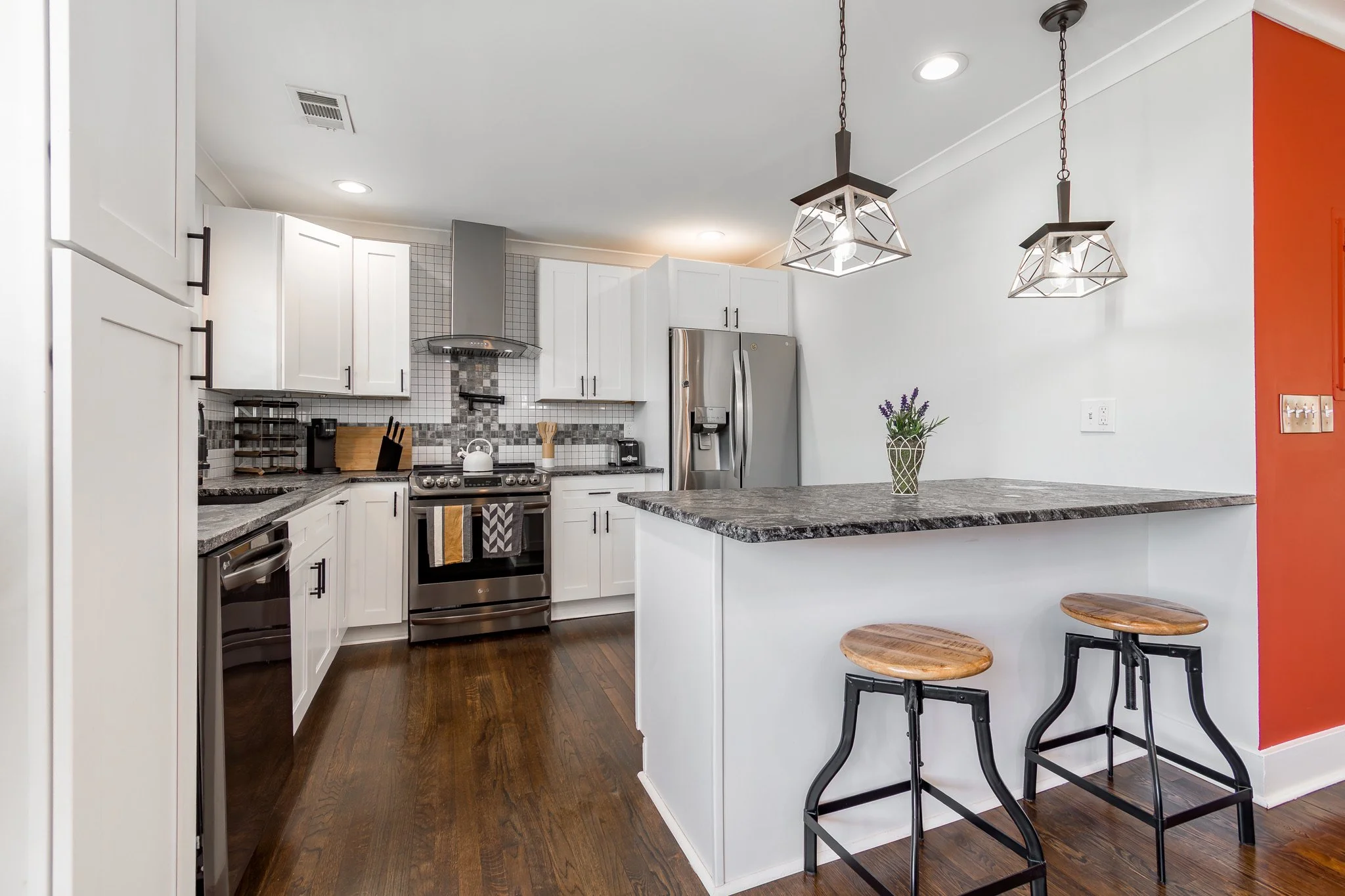 Modern kitchen with white cabinets, stainless steel appliances, dark granite countertops, a small kitchen island with two bar stools, a potted lavender plant, red accent wall, and wood flooring.