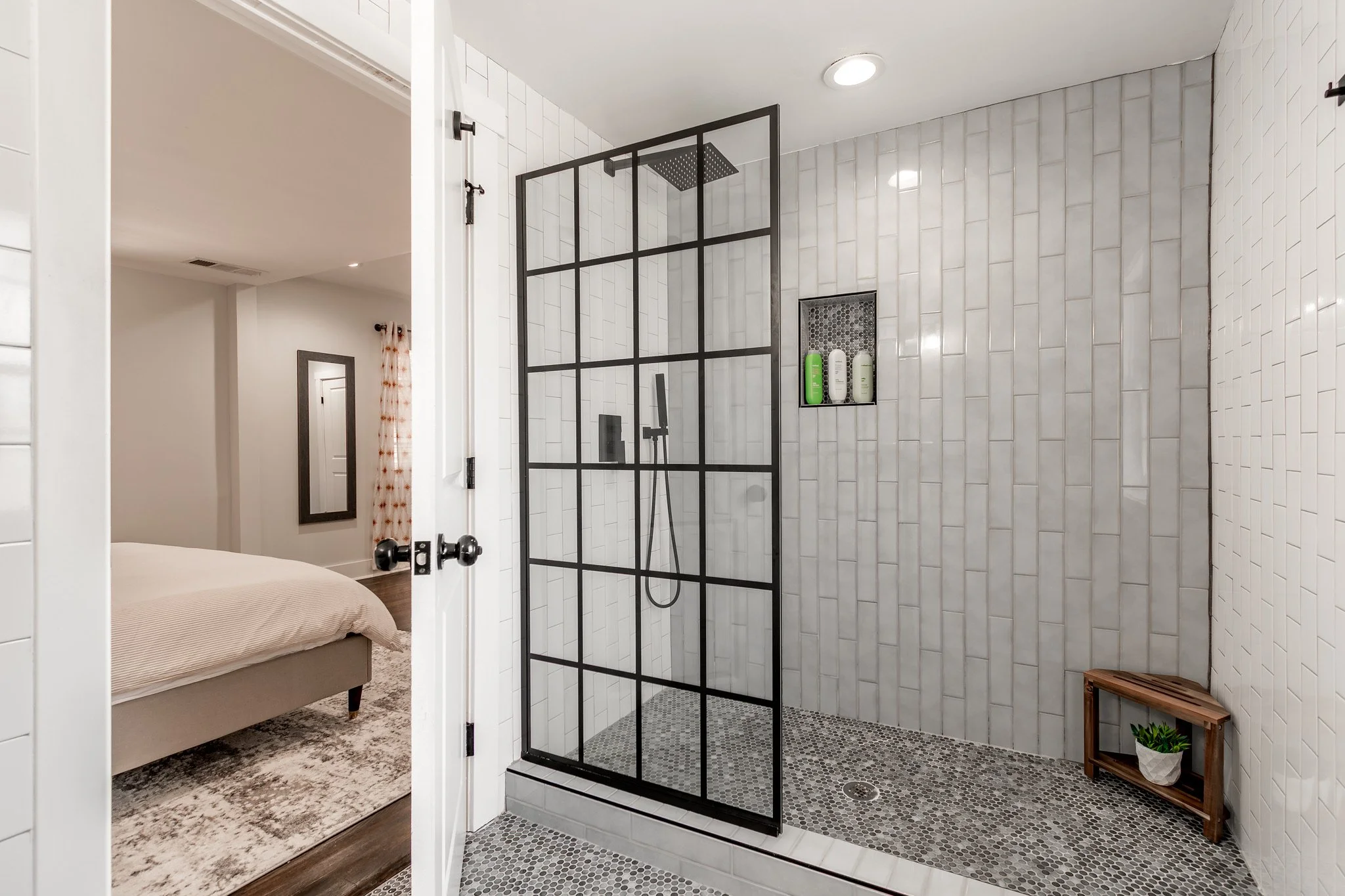Modern walk-in shower with black-framed glass door, white tile walls, small pebble floor, built-in niche with shampoo bottles, and a small wooden shelf with a plant inside.