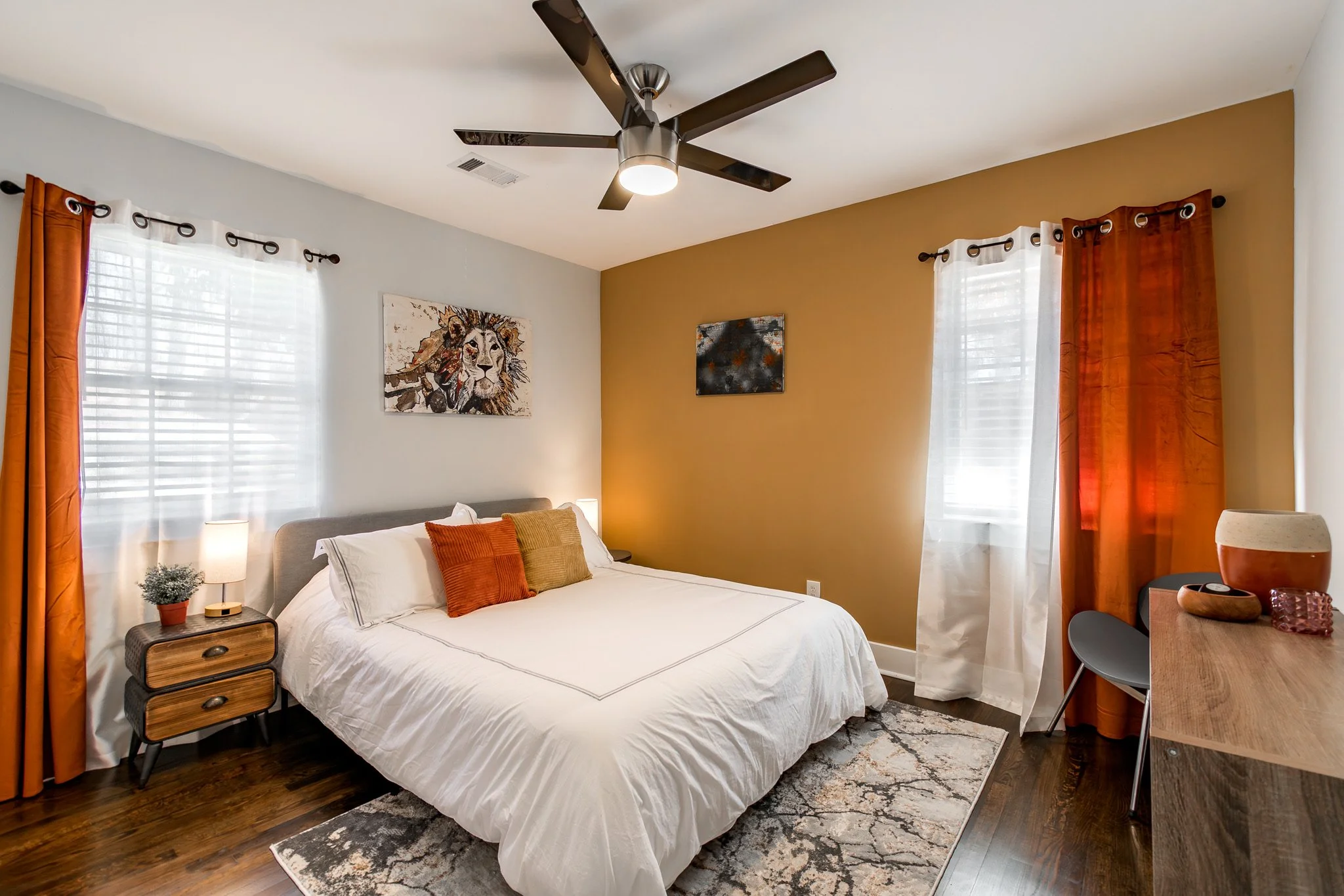 Modern bedroom with white bed, orange and beige pillows, wooden nightstand, potted plant, artwork of a lion and abstract painting, hardwood floor, and windows with orange and white curtains under ceiling fan.