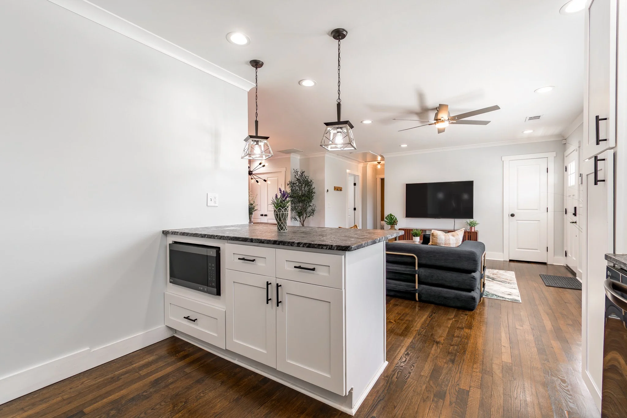Open-concept kitchen and living room with white cabinets, dark granite countertops, hardwood floors, black pendant lights, ceiling fan, wall-mounted TV, and a dark sofa with pillows.