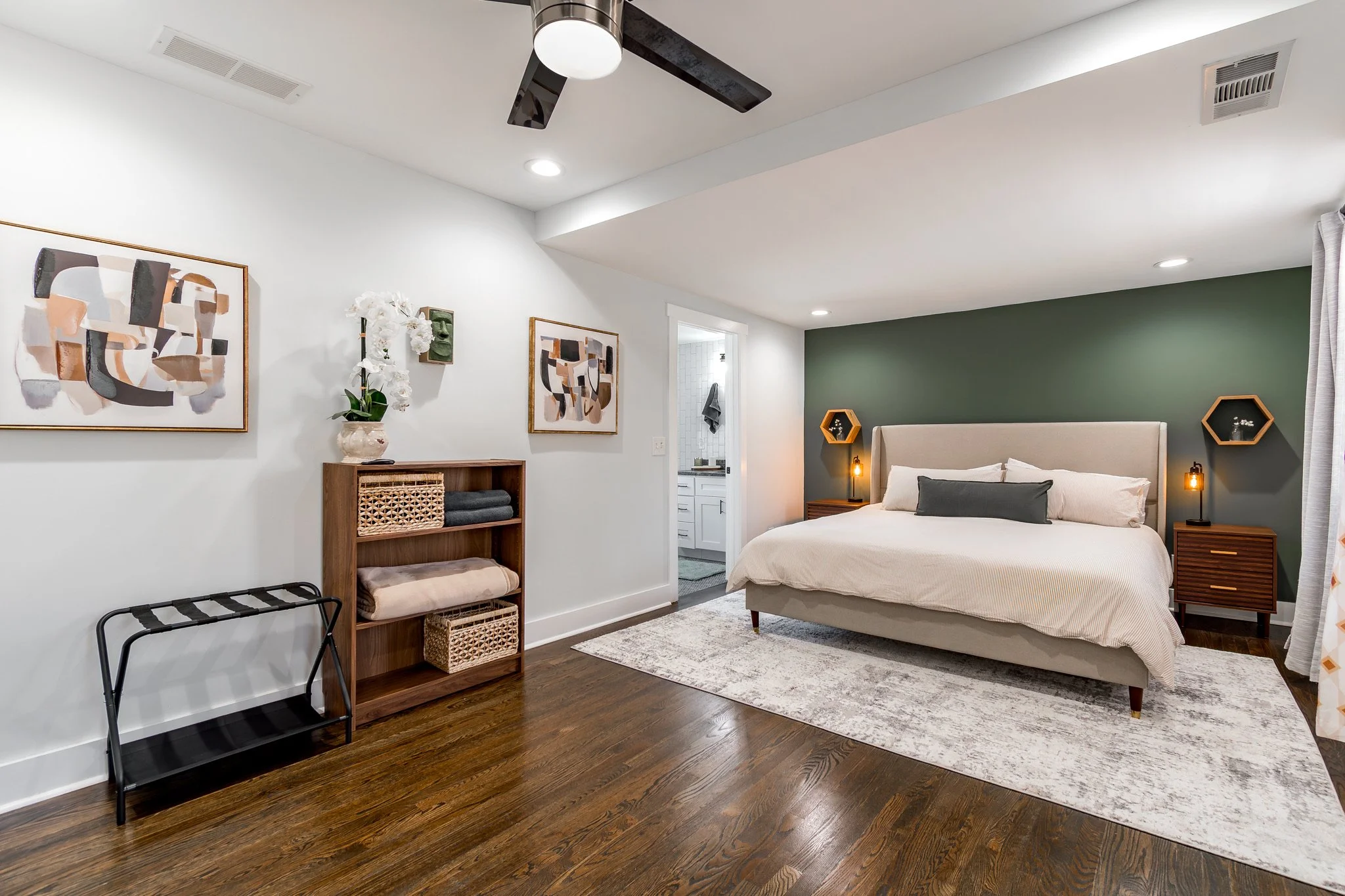 Modern bedroom with white walls, a green accent wall, a large bed with white bedding and a dark pillow, wooden nightstands with bedside lamps, a wooden bookshelf, abstract art on the walls, and a ceiling fan.