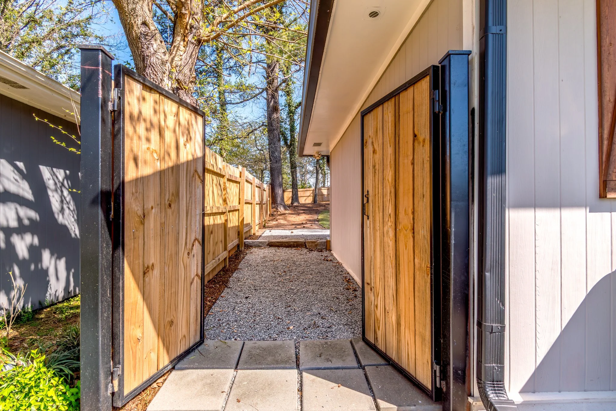 Side yard gate with wooden panels open, leading to a backyard with trees and a gravel pathway, next to a white house with blue trim.