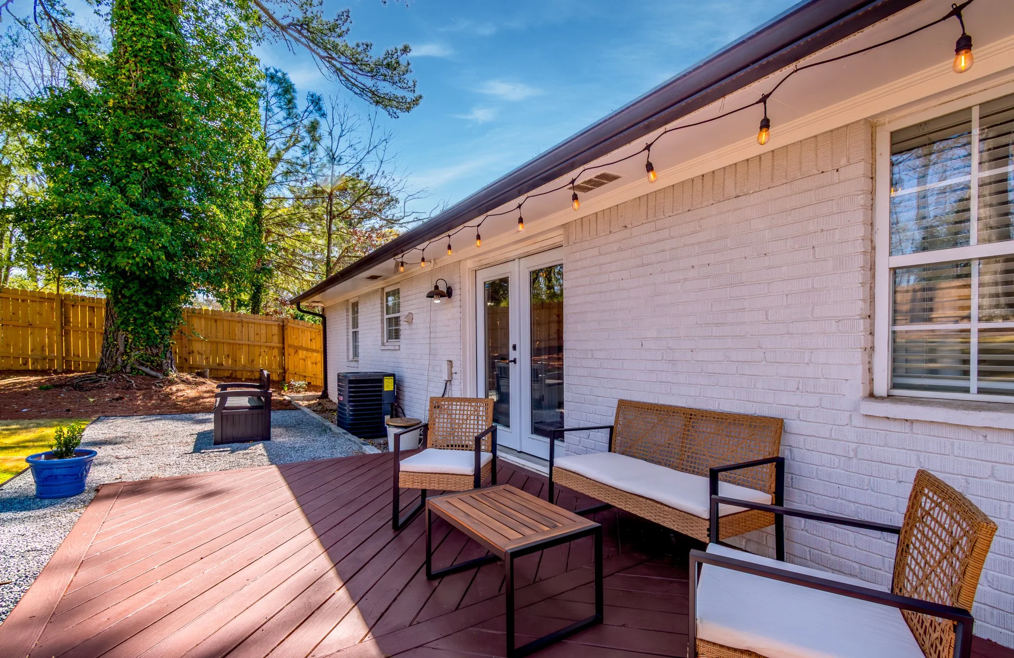 Backyard patio with outdoor furniture, string lights, a large tree, and a white brick house with a window and a sliding door.