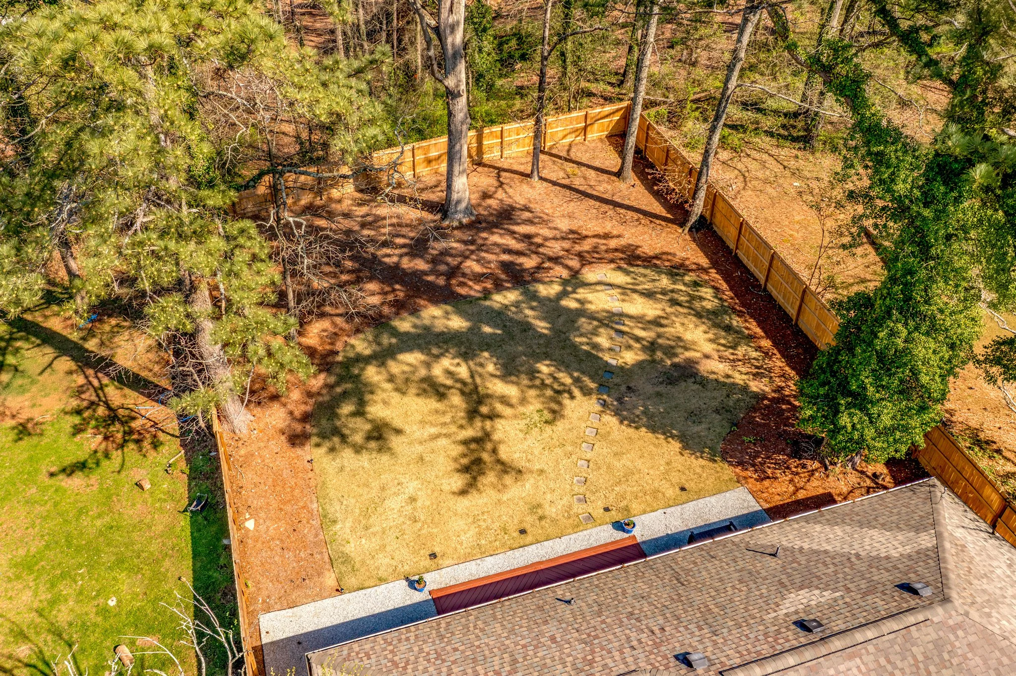 An aerial view of a backyard with a wooden fence, several trees, a lawn area with stepping stones, and part of a house with a shingled roof.