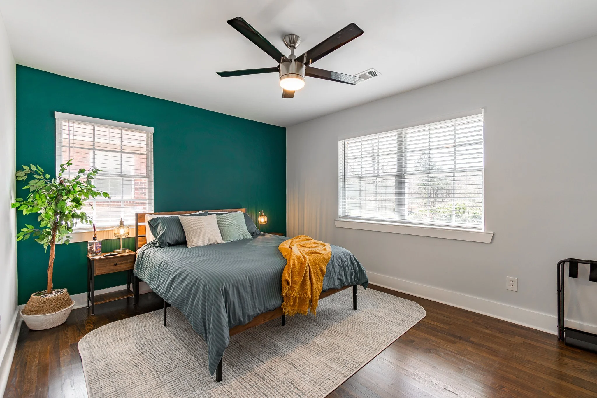 A bedroom with a dark green accent wall behind a bed with gray bedding and multiple pillows. A small wooden nightstand with lamps and decor on each side of the bed. Large windows with blinds letting in natural light, and a ceiling fan with black blad
