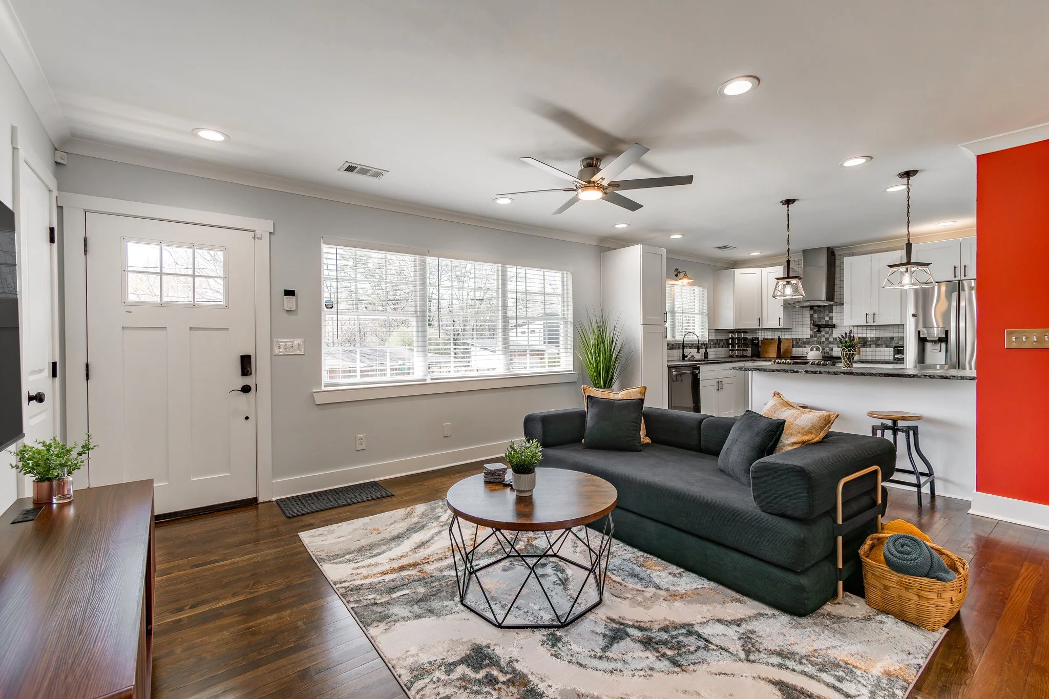 Modern living room with dark sofa, round coffee table, large windows, and open kitchen with white cabinets and stainless steel appliances.