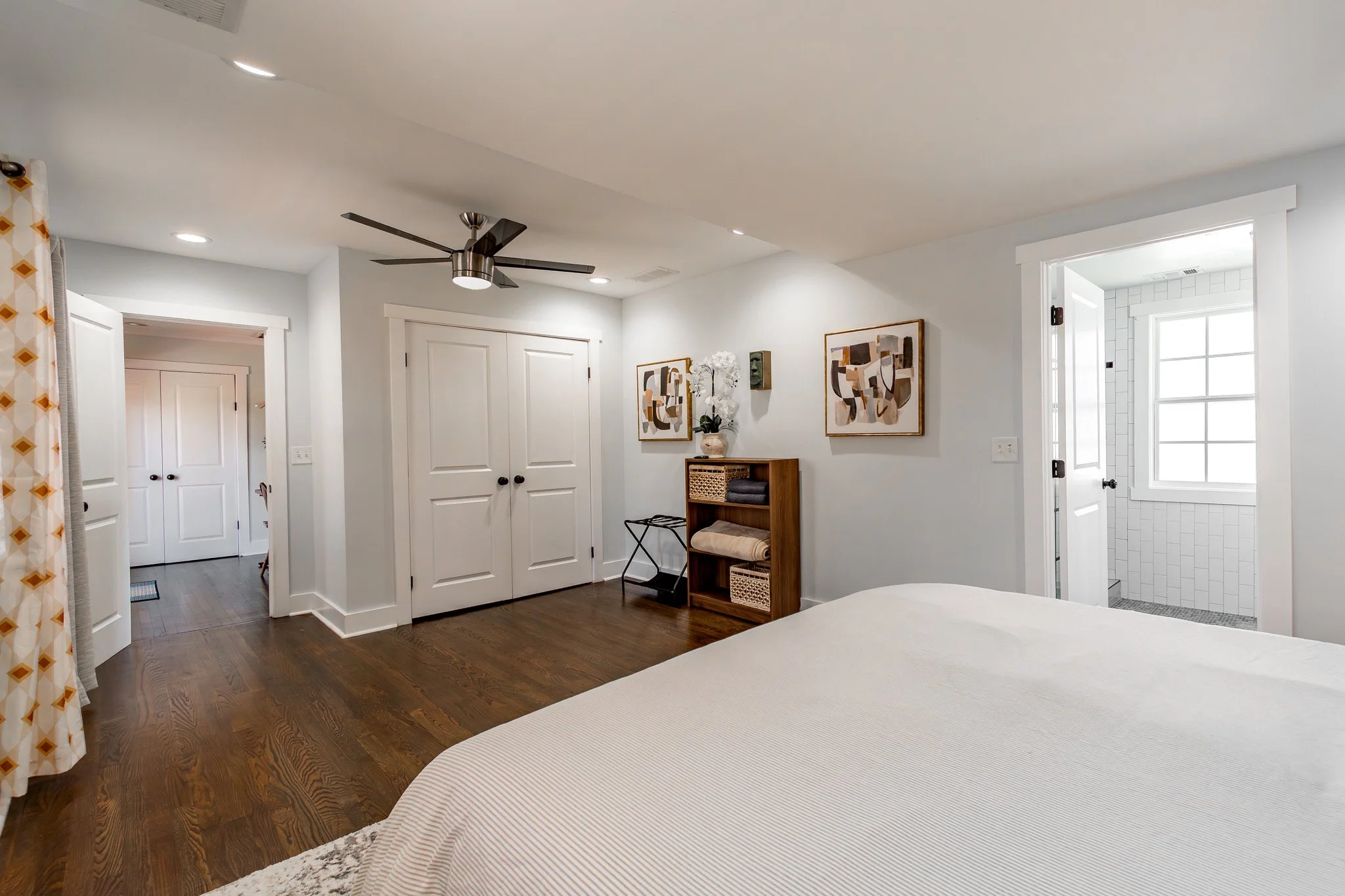 A bedroom with a white bed, wooden floor, white closet doors, abstract art on the walls, a ceiling fan, and a small open bathroom with a window.