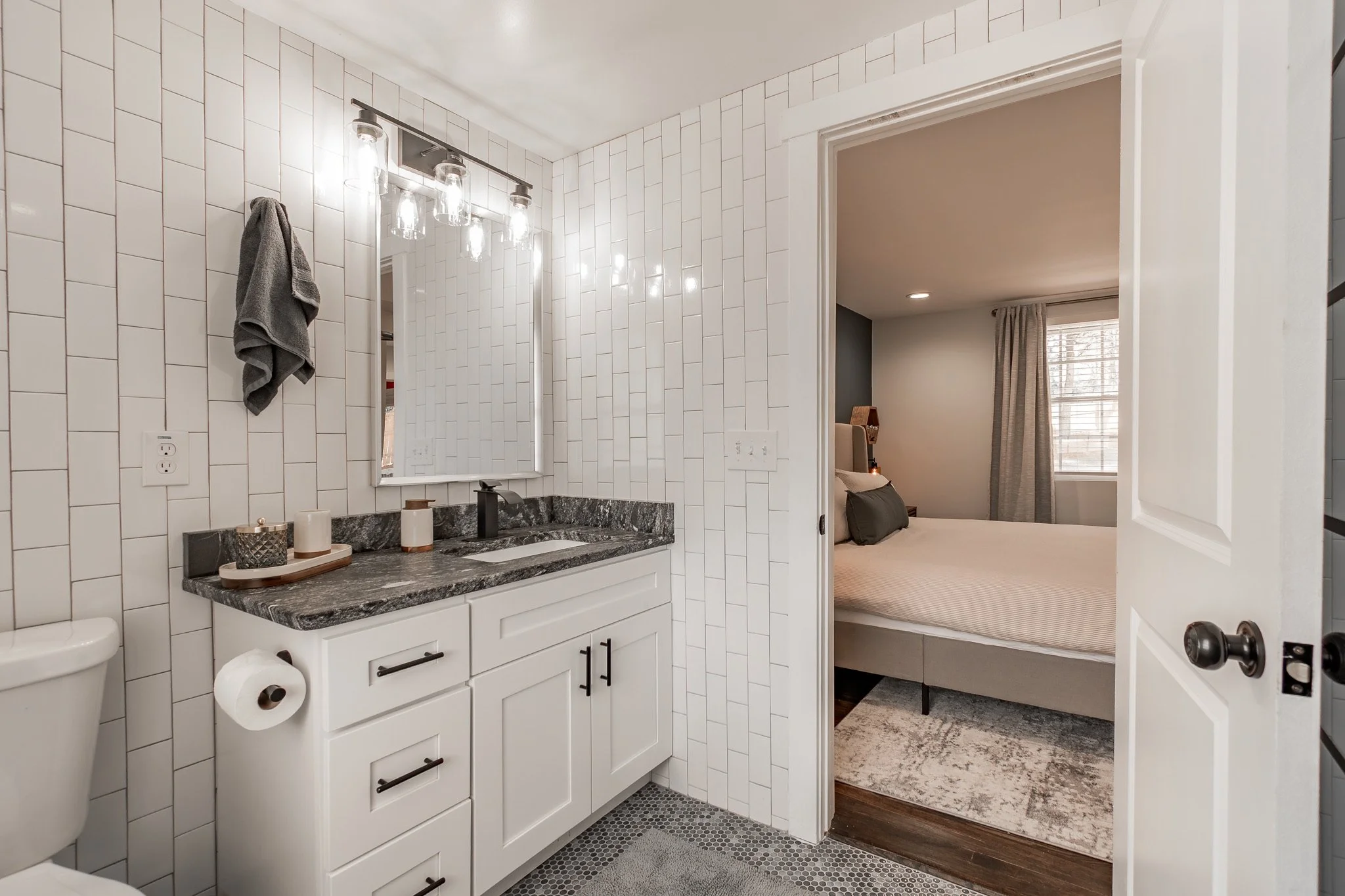 Modern bathroom with white tile walls, black granite countertop, mirror, wall-mounted light fixture, gray towel on wall, toilet, and doorway leading to a bedroom with a bed, window, curtains, and bedside lamp.