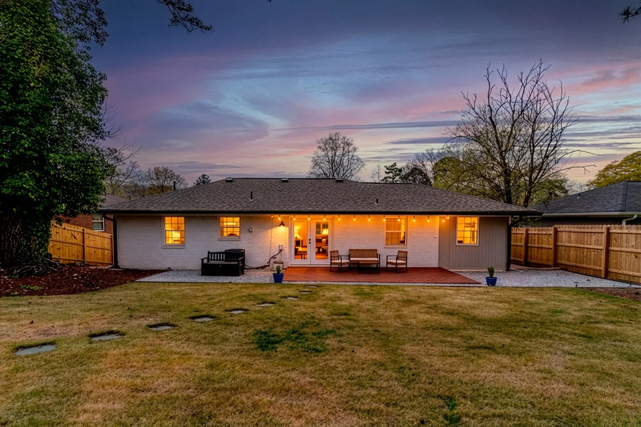 Backyard of a house during sunset with a lawn, wooden fence, and outdoor seating area with string lights.