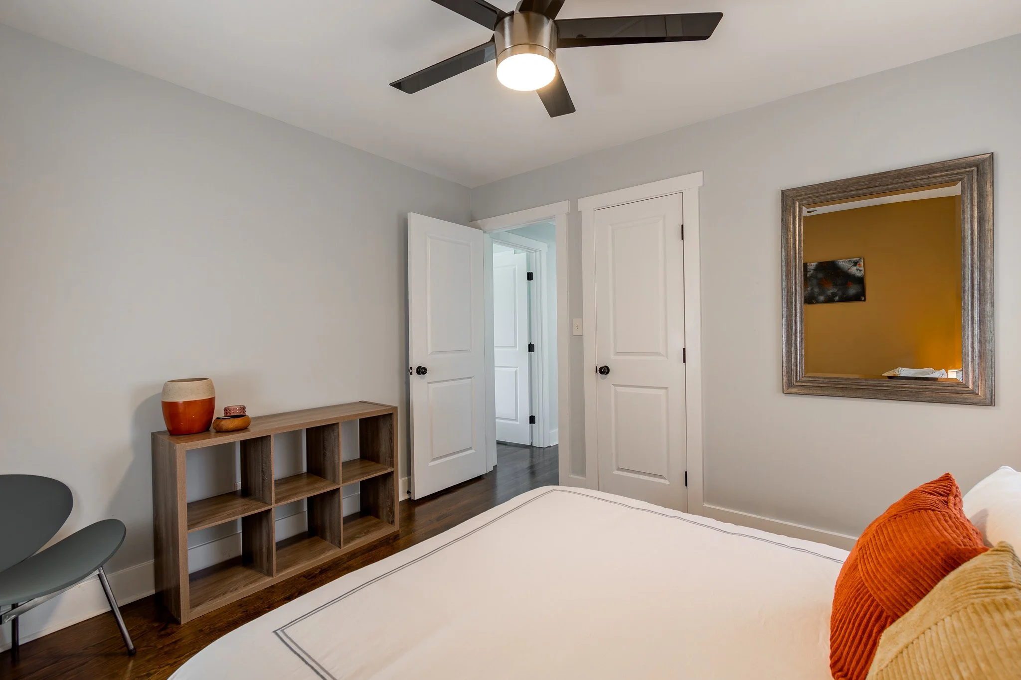 A bedroom with white walls, a ceiling fan, a mirror on the wall, a wooden shelf, and a bed with orange pillows.