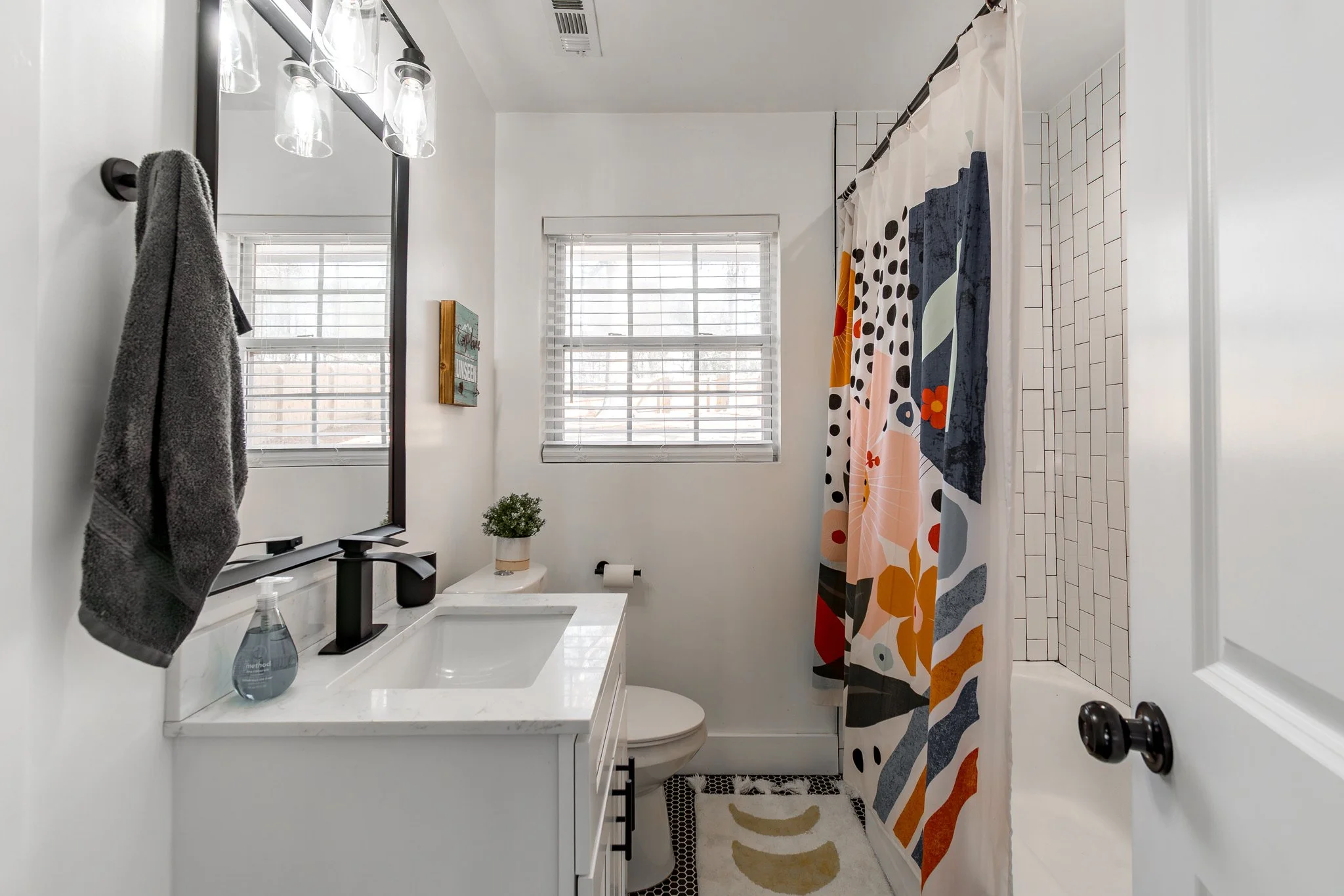 A small bathroom with a white vanity, black faucet, and a mirror. There are two windows with blinds, a decorative plant, and colorful patterned shower curtain. Towel hanging on wall, toilet paper on holder, and light fixture with glass shades.