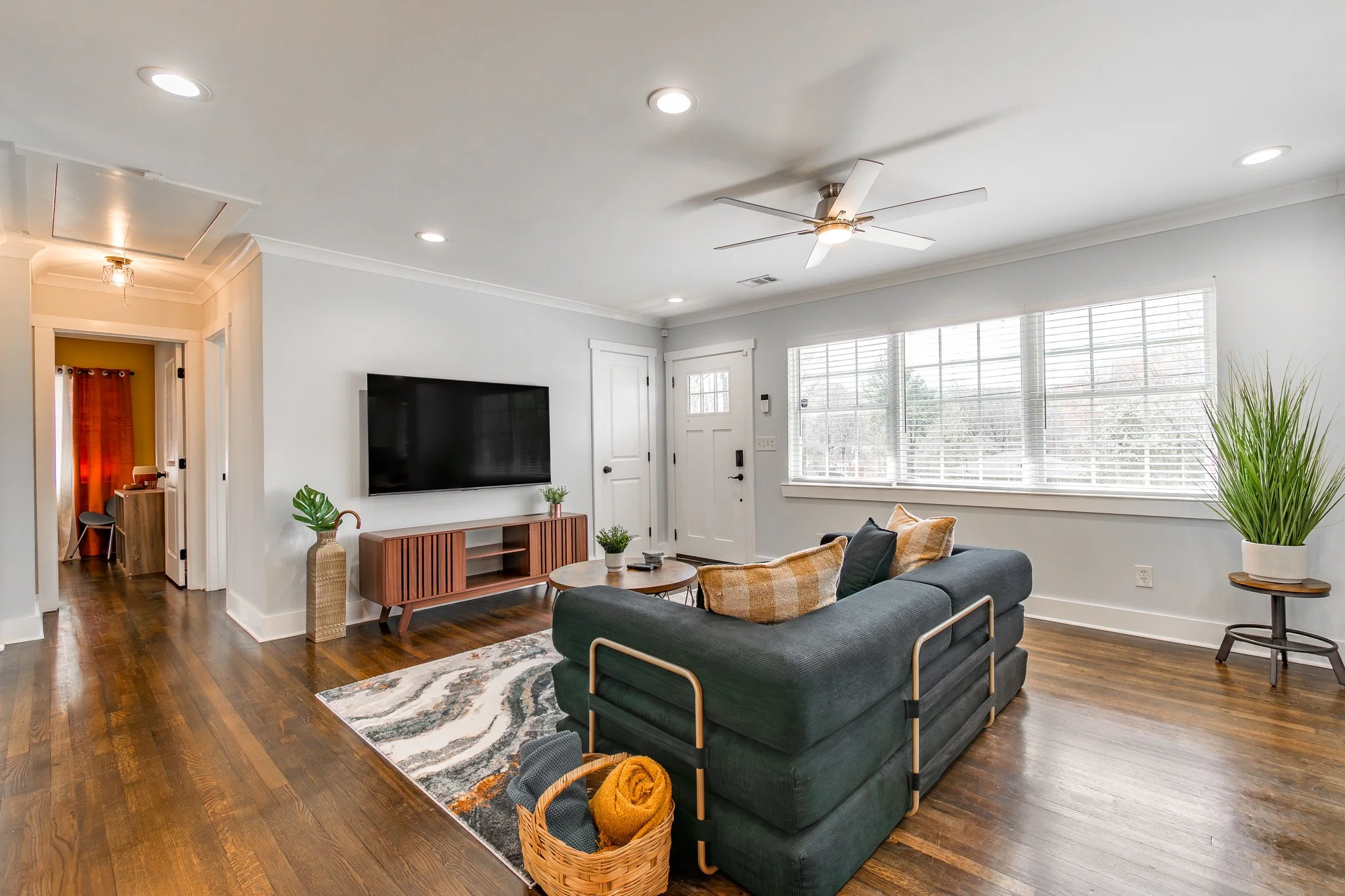 Living room with white walls, wooden floor, large window with blinds, black sofa with pillows, round coffee table, TV mounted on wall, tall green plant, and wicker basket on the floor.