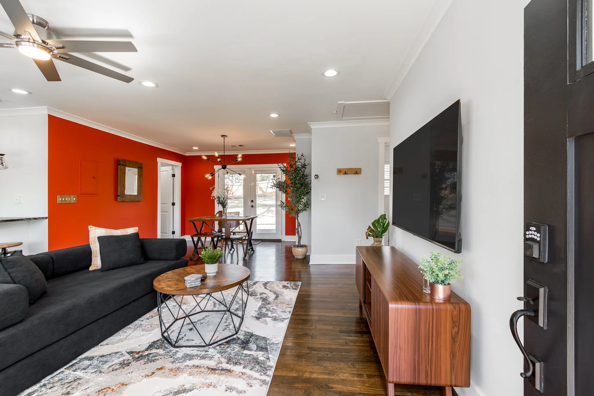 Open-concept living room and dining area with a vibrant orange accent wall, a black sofa, a modern wood TV console, a large wall-mounted TV, and a contemporary dining table with chairs near a set of glass doors leading outside.