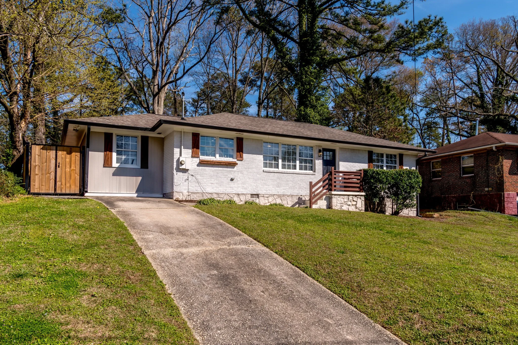 A single-story house with a light brick exterior, wooden shutters, and a small front porch with wooden stairs. The house is set on a grassy hill with a concrete driveway leading up to it. There are tall trees and a clear blue sky in the background.
