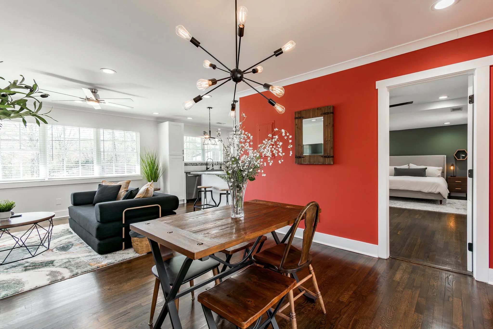 Open-concept living room and dining area with a red accent wall, a wooden dining table with chairs, a black sofa, large windows with white blinds, and a view into a bedroom with green wall.