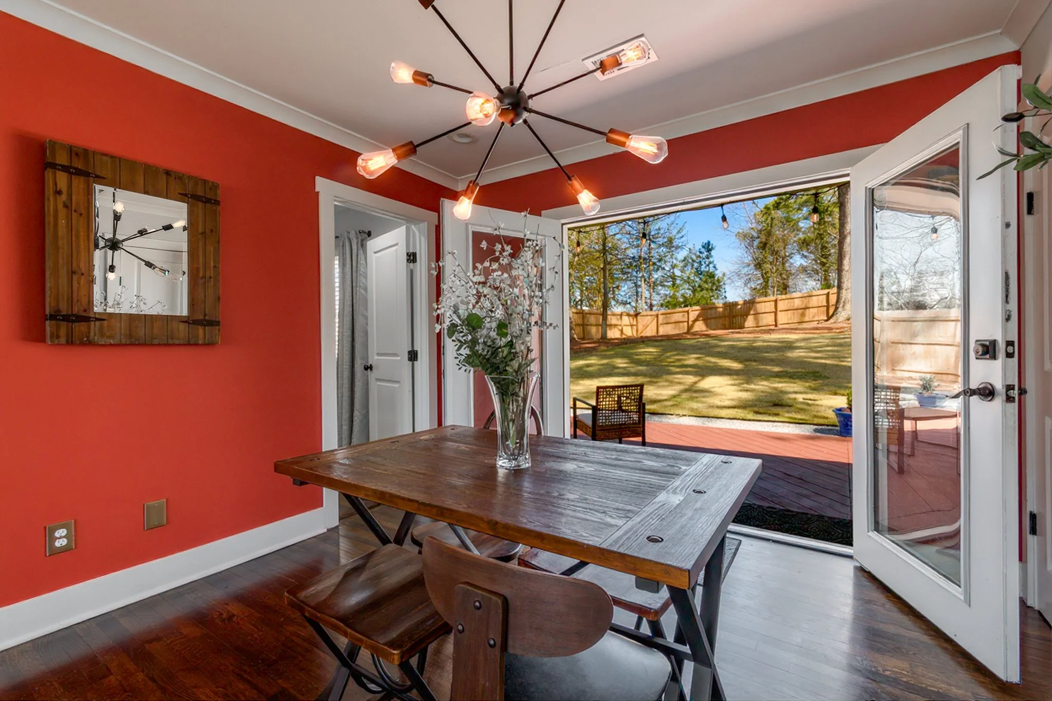 Interior view of a dining room with red walls, a wooden table, a glass vase with flowers, and an open sliding glass door leading to a backyard.