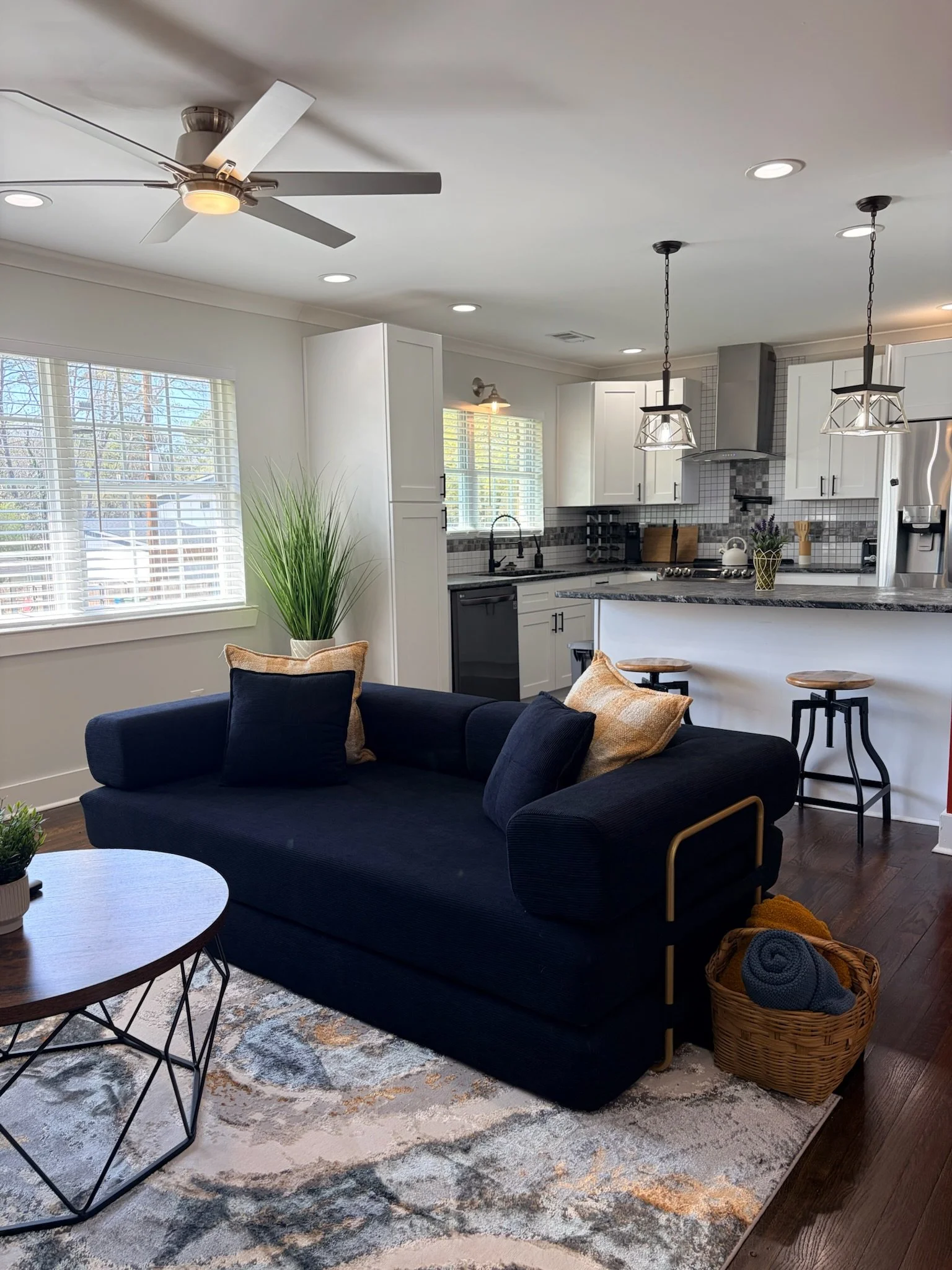 Living room with dark sofa, decorative pillows, wooden side table, area rug, kitchen with white cabinets, pendant lighting, and a ceiling fan.